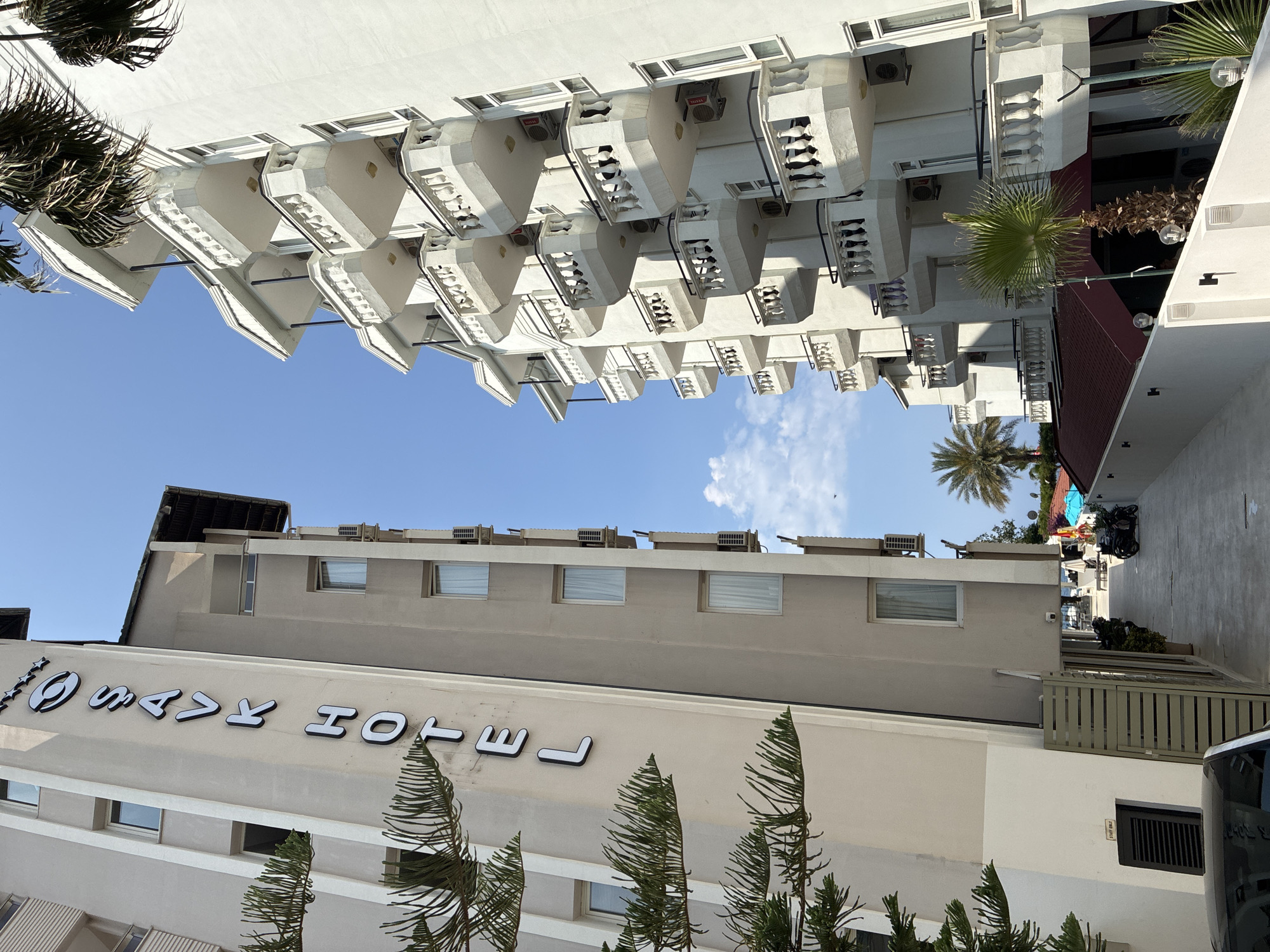 The image depicts two hotel buildings in Alanya, Turkey, under a clear blue sky. The building on the left is light beige with the vertical sign "SAVK HOTEL" in white lettering. It has several floors with windows and some trees in the foreground. The building on the right is white and features multiple balconies with ornate railings. In the background, there are palm trees and a glimpse of the sea. The scene suggests a sunny day in a resort town. There are no people clearly visible.