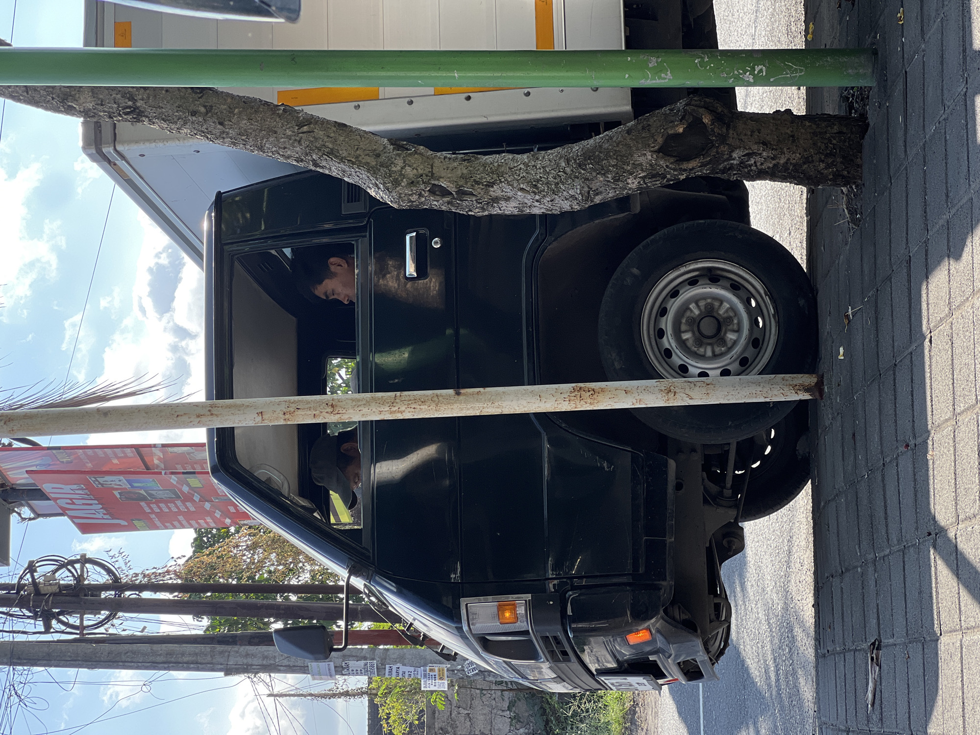 This image depicts a black commercial truck parked on the side of a street in Denpasar, Indonesia. The truck is dark green and has a silver cargo box attached to the rear. Two men are visible inside the truck's cab. One man, wearing a dark baseball cap, is looking down at something on his lap. The second man, with short black hair, is looking out the passenger side window. A tree with rough bark grows closely to the side of the truck, with a green utility pole standing next to it. The street is paved with concrete tiles, and there are power lines and a red billboard visible in the background. The lighting suggests it is daytime.