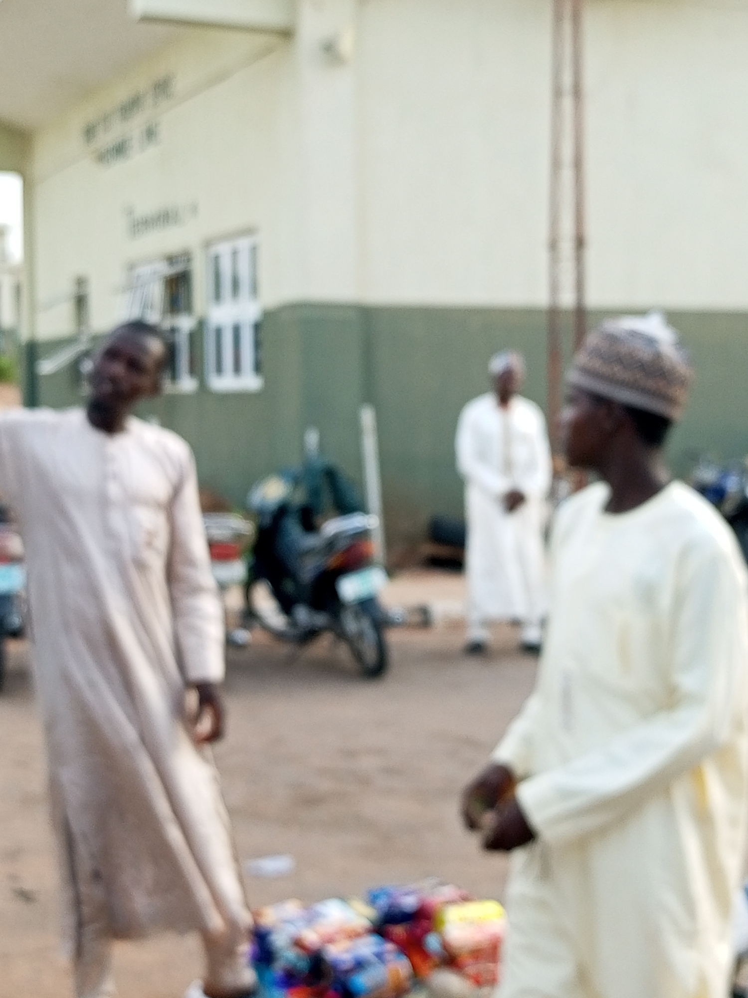 In the city of Gombe, Nigeria, a shallow depth of field photograph captures three men in traditional attire. In the foreground, slightly off-center to the right, a man in a light yellow kaftan and patterned cap is looking towards the right of the frame. His hands are clasped in front of him. To the left of the frame, a man in a tan kaftan is blurred and appears to be gesturing with his left hand. Behind these two figures, in the mid-ground, a third man in a white kaftan stands with his hands at his sides. The men are standing on a dirt or gravel surface. In the background, a light-colored building with green trim is visible. The building has windows and appears to be a public or commercial structure. To the left of the building, there is a glimpse of what might be a street or walkway. Between the two foreground men, a motorcycle is parked. Piles of colorful items, possibly goods for sale, are visible on the ground in the lower right portion of the image. The overall scene suggests an outdoor marketplace or a gathering point near a building.