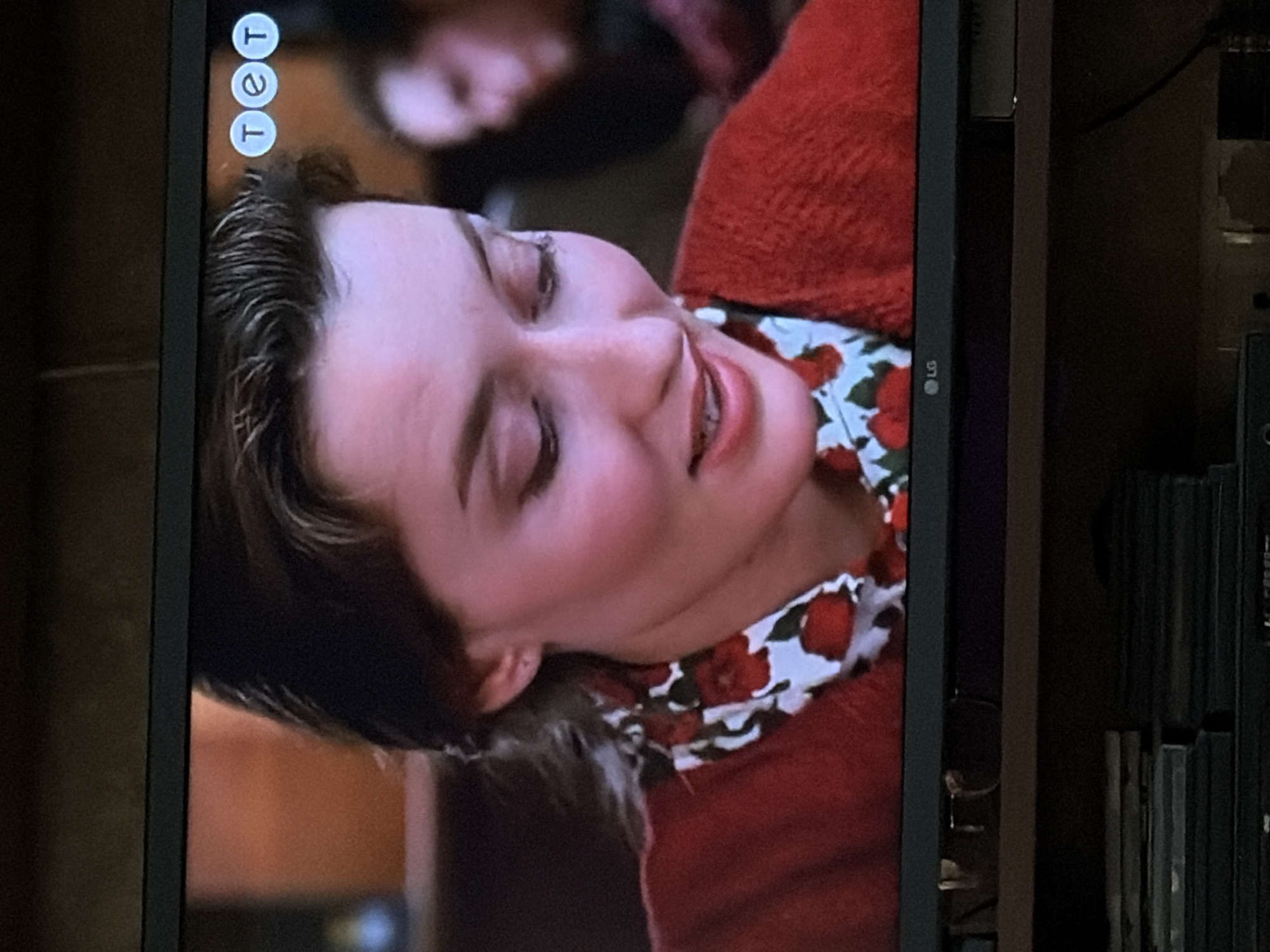 A woman with dark hair pulled back, wearing a red knitted sweater over a white shirt with a red floral print, is shown from the chest up, looking down and to the left with a slight smile. She has fair skin and is wearing subtle makeup. In the upper right corner of the frame, three white circles with blue letters inside form the logo "tet." The background is blurred, showing warm-toned elements and a hint of another person's face on the right. Below the screen, a black LG logo is visible on the television frame, and stacked media cases are visible on a shelf underneath. The scene appears to be indoors, likely in a living room or studio setting.