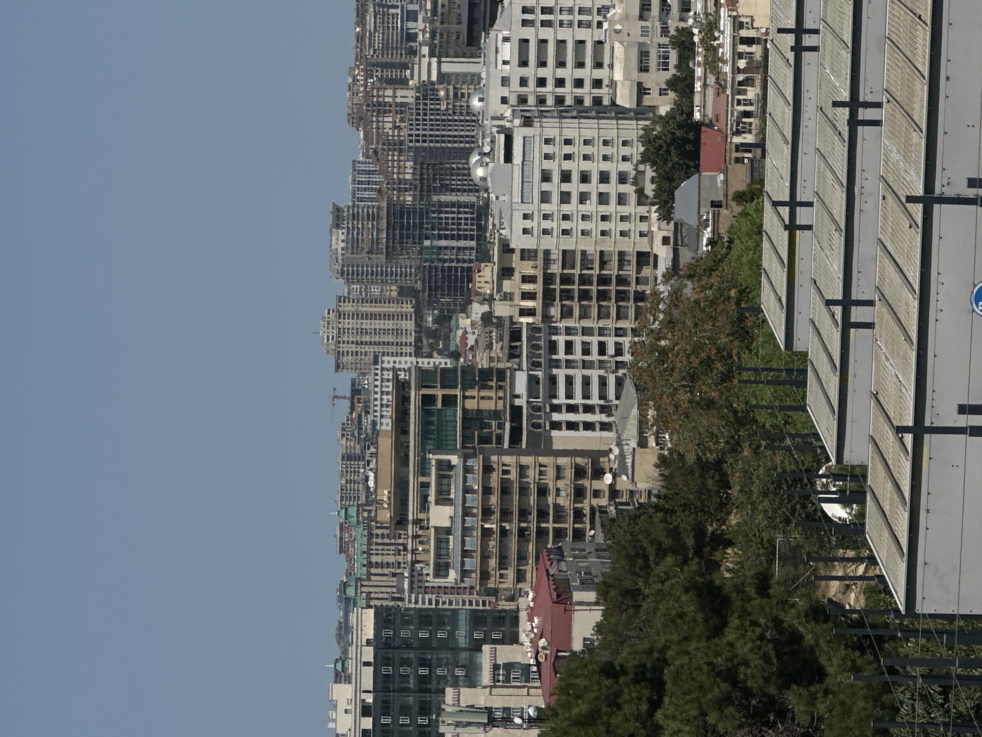 This is a high-angle, full-frame, daylight shot of a cityscape in Sabail Raion, Azerbaijan. The background consists of a clear, pale blue sky. In the middle ground, a dense collection of multi-story buildings dominates the view, showcasing a variety of architectural styles and ages. Many buildings appear to be residential or office towers, with numerous windows visible. Some structures are partially under construction, with exposed frameworks.
In the foreground, there are several layers of structures. The most prominent are rows of rectangular, light-colored, roof-like structures with corrugated metal or plastic sheeting, supported by dark metal poles. These appear to be utilitarian, possibly covering outdoor equipment or storage. Between these foreground structures and the main cityscape, there are dense areas of dark green trees, and some lighter green foliage, with visible trunks and branches. Fencing and vertical posts are also present in this mid-foreground area.
The overall impression is one of urban development with a mix of completed buildings, ongoing construction, and natural elements.