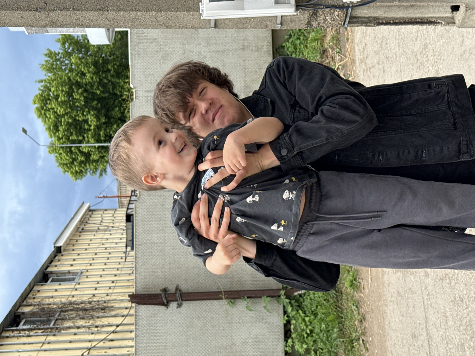 In Chaikovsky, Russia, a young person in a black denim jacket holds a small child wearing a black shirt with a Snoopy pattern. The child is looking to their left with a smile, while the young person gazes forward with a slight smile. They are standing outdoors, with a grey concrete fence behind them. To the left, a weathered yellow building with visible wooden studs and windows can be seen. Further in the background, a vibrant green tree stands against a cloudy sky, and a street light is visible. A concrete pillar with electrical equipment is on the right side of the frame. The ground appears to be a mix of gravel and asphalt.