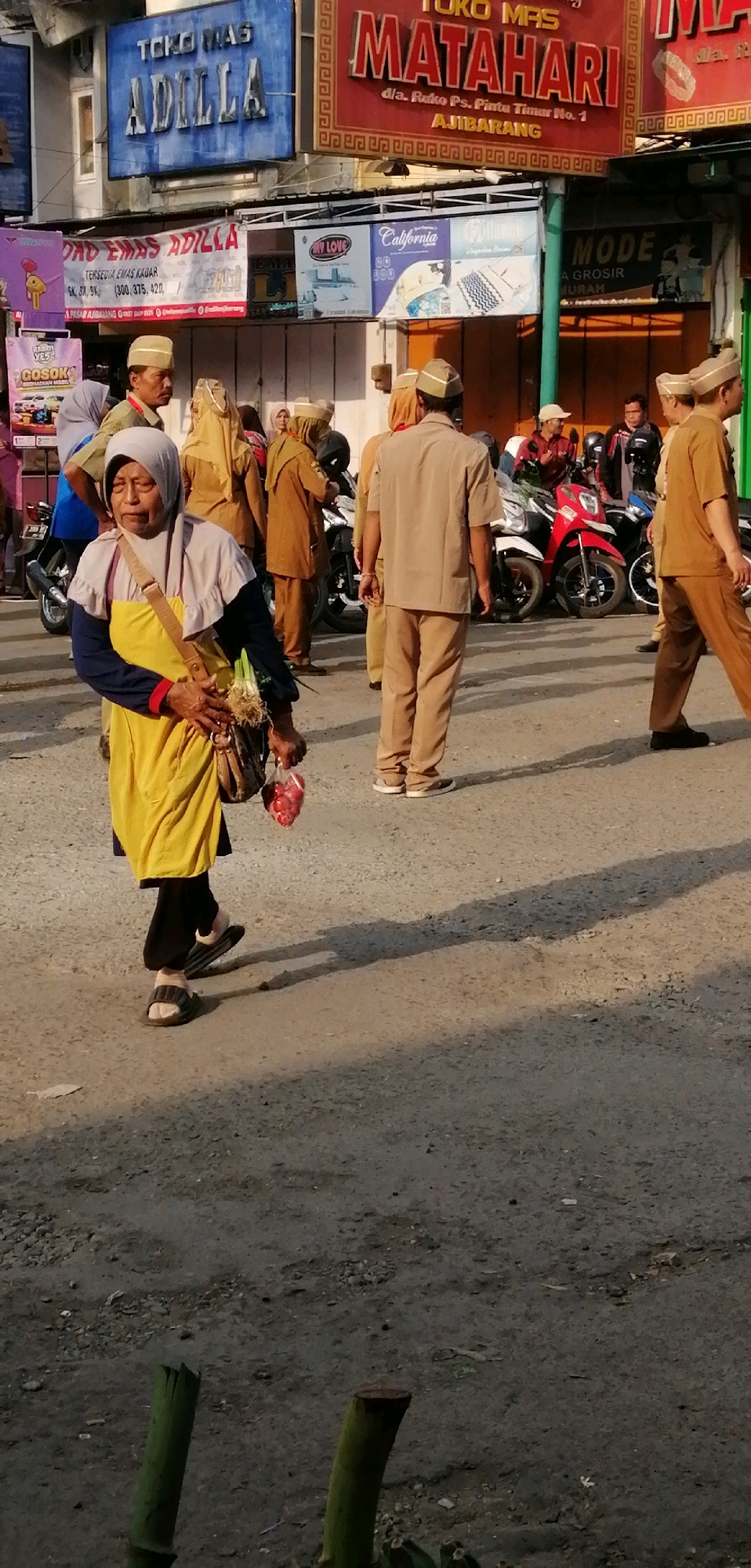The image depicts a street scene in Ajibarang, Indonesia, likely during daylight hours given the shadows. In the foreground, a woman wearing a hijab, a yellow apron, and dark pants walks towards the left. She carries a bag over her shoulder and holds a plastic bag containing red items, possibly meat, and a bunch of green vegetables in her left hand. She is wearing sandals. Behind her, several individuals in matching tan uniforms with similar hats are standing and walking. Some are facing away from the camera, while others are partially visible from the side. To the right of the woman, a man in a tan uniform walks away from the camera. In the background, there are signs for shops, including "TOKO MAS ADILLA" and "TOKO MAS MATAHARI." Several motorcycles are parked in the background. The ground appears to be a dusty, paved surface. The overall atmosphere suggests a bustling market or commercial area.