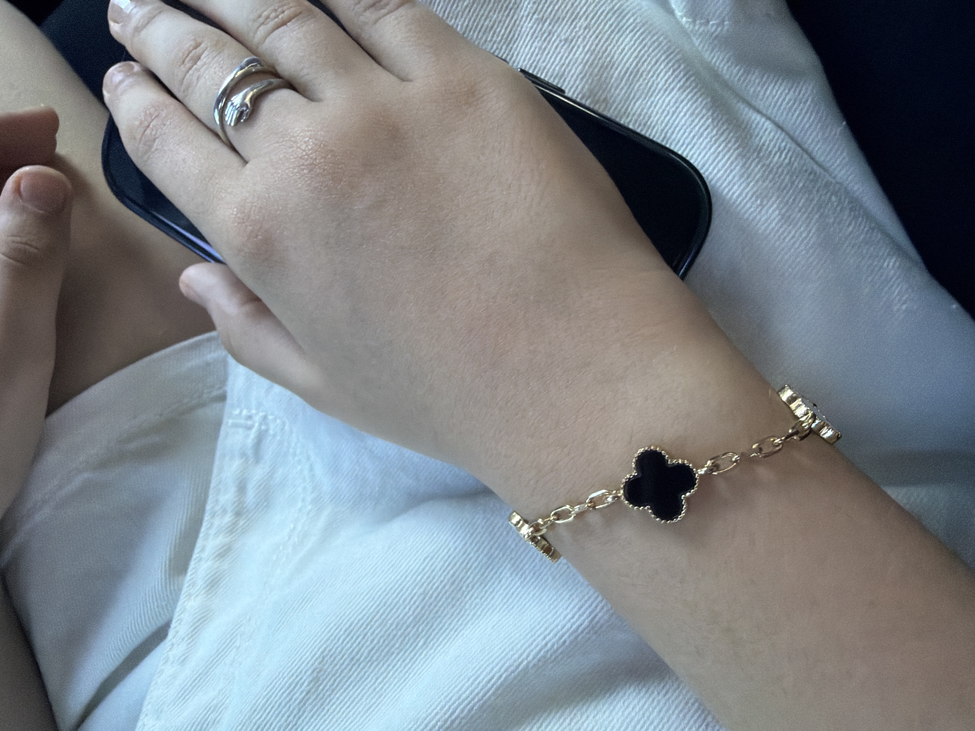 A close-up shot focuses on a person's left arm and hand, adorned with jewelry and resting on a black smartphone. A gold bracelet with black clover-shaped charms is visible on the wrist. A silver ring, designed to look like two arms hugging, is worn on the index finger. The person is wearing white fabric, possibly clothing. The image does not contain any information or visual cues to determine the location in Yerevan, Armenia.