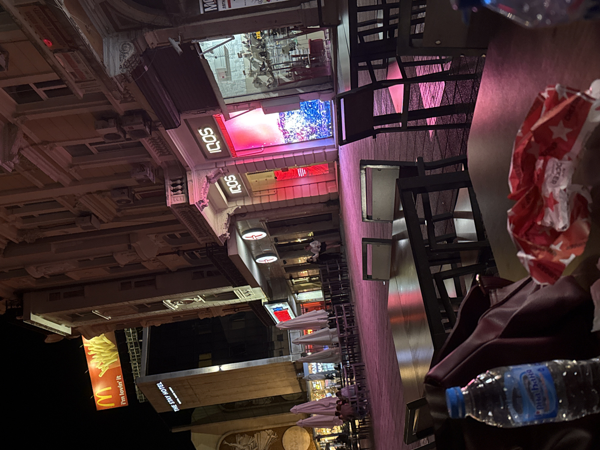 The image depicts an evening street scene in Plovdiv, Bulgaria, showcasing a vibrant urban environment with shops, restaurants, and outdoor seating areas. The perspective is slightly elevated, looking down a street lined with buildings illuminated by numerous neon signs against a dark night sky.
In the foreground, a dark table holds a plastic bottle of "Горна баня" (Gorna Banya) water, a burgundy bag, and a crumpled red and white wrapper. Beyond this, an outdoor dining area features dark tables and chairs, some covered by white, folded umbrellas. Several people are visible enjoying the evening at these tables.
The buildings lining the street display various illuminated signs. Multiple "IQOS" signs are prominent, along with a large, colorful LED screen. Further down, a large McDonald's sign with french fries and the "i'm lovin' it" slogan is visible, as is a sign for "THE STAY HOTEL." The architectural details of the buildings suggest historical significance.
The time of day is clearly night, with artificial lights providing the primary illumination. The scene appears lively and bustling, characteristic of a commercial street or pedestrian zone. Other partially visible text includes "MO", "лятн", and "HA" on various signs.