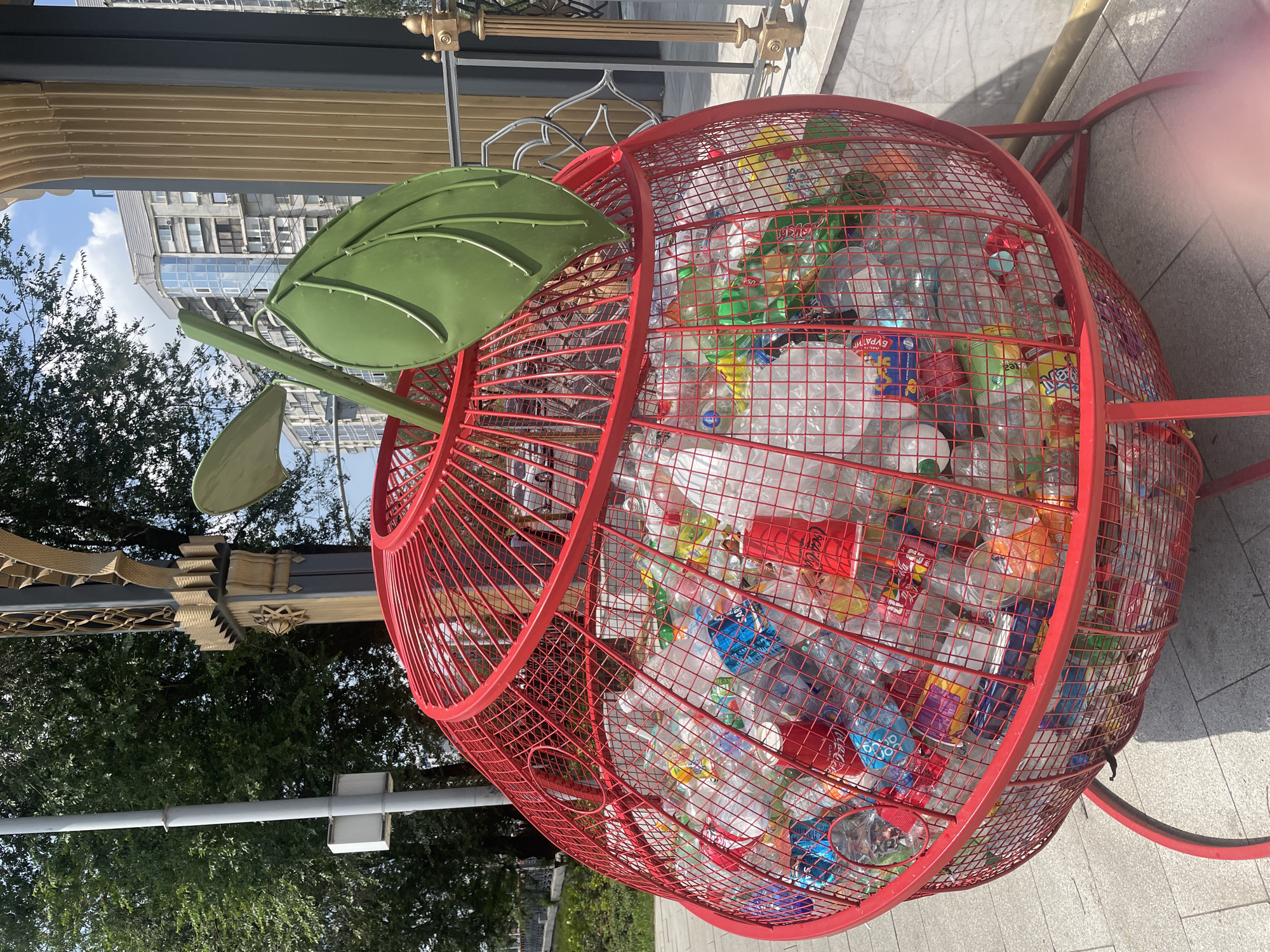 The image shows a large, red, apple-shaped bin filled with plastic bottles. The bin is made of a red metal mesh and has a green leaf and stem attached to the top, further enhancing its apple appearance. The bin is situated outdoors on a paved surface, likely a sidewalk or public area. In the background, there are trees, buildings, and a clear blue sky with some clouds, suggesting it is daytime and the weather is fair. The bin appears to be a creative and environmentally conscious way to collect recyclable plastic waste. There are no people visible in the image. The primary object of focus is the apple-shaped recycling bin overflowing with various plastic bottles, some with visible labels. The overall scene is bright and suggests an urban or public setting in Almaty, Kazakhstan.