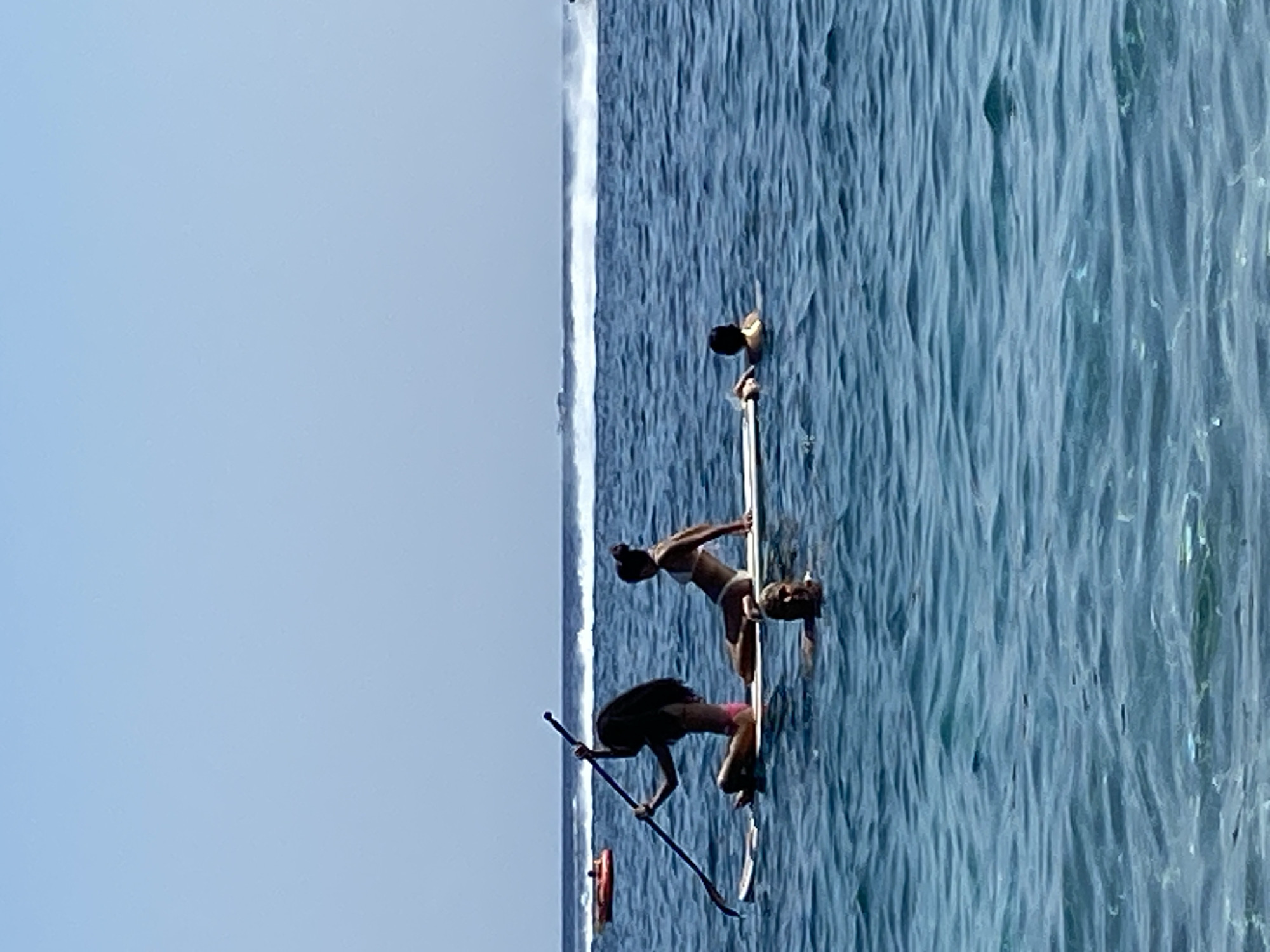 The image depicts a serene scene in the waters of Prijevor, Montenegro. In the foreground, the clear blue water ripples under a bright sky. A white paddleboard is the central object, with three individuals on or near it. On the paddleboard, a person with dark hair sits cross-legged, holding a paddle. Next to them, another person in a white bikini is reclining. In the water beside the paddleboard, a person's head is visible, seemingly swimming. In the background, a red inflatable tube is visible on the left, and on the right, a wake suggests a fast-moving boat or jet ski in the distance. The horizon is marked by a clear blue sky meeting the ocean, with some distant white streaks on the water.