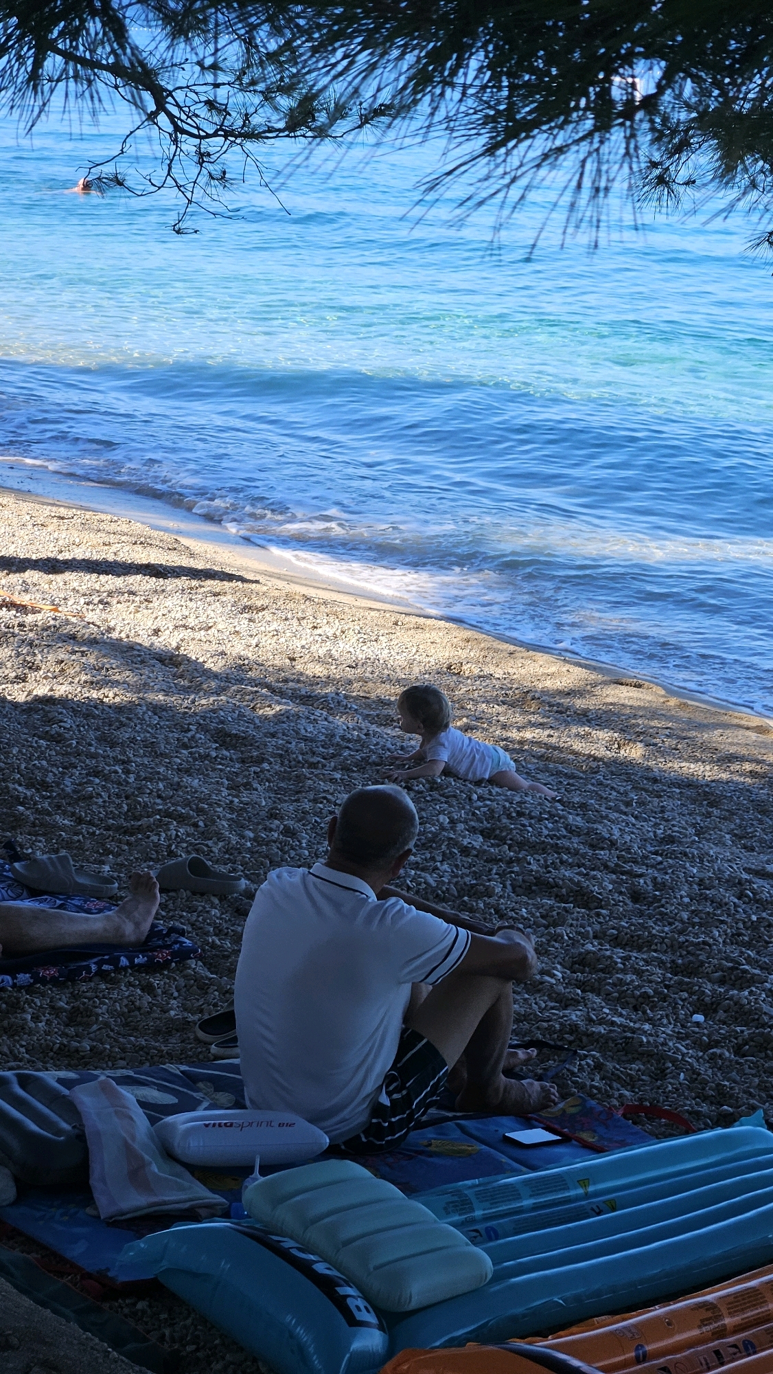 The image depicts a pebble beach in Općina Tučepi, Croatia, with the turquoise Adriatic Sea in the background. A person, viewed from behind, sits on a blue inflatable mattress on the shore. Beside them, a baby is crawling on the pebbles, facing the water. In the distance, a person is swimming or wading in the shallow, clear water. Pine branches hang over the scene from above, casting shadows on the beach. Various other beach items, including towels and inflatable objects, are spread out on the ground.