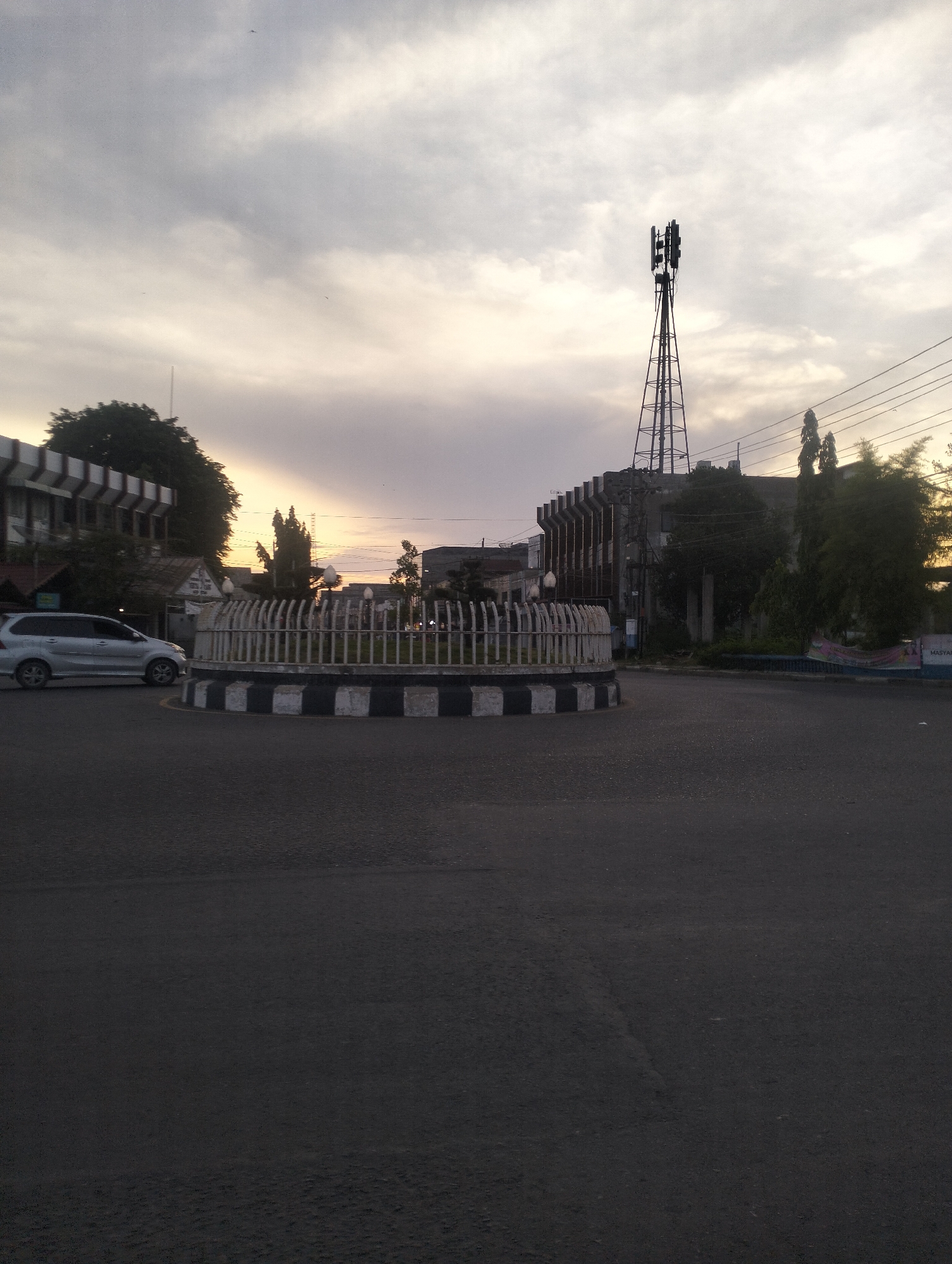 The image depicts an urban street scene in Lhokseumawe, Indonesia, captured during either sunrise or sunset. The central feature is a circular roundabout with a distinctive white and black striped curb, enclosed by white decorative fencing and containing sparse greenery. A silver multi-purpose vehicle, resembling a Toyota Avanza, is seen on the left, navigating the roundabout.
In the background, a multi-story building with a unique facade is visible on the left, partially obscured by trees. On the right, a tall communication tower equipped with antennas stands prominently against the sky, behind other buildings and trees. Overhead power lines are visible in the upper right quadrant.
The sky is covered with clouds, allowing diffused light to filter through, with a warm, golden-orange hue visible near the horizon, indicative of either early morning or late afternoon. The overall lighting is dim, casting long shadows and lending a calm atmosphere to the scene. A banner on the far right is partially visible, showing the text "MASYAR...", likely part of an Indonesian word such as "Masyarakat" (community). No individuals are explicitly visible in the scene.