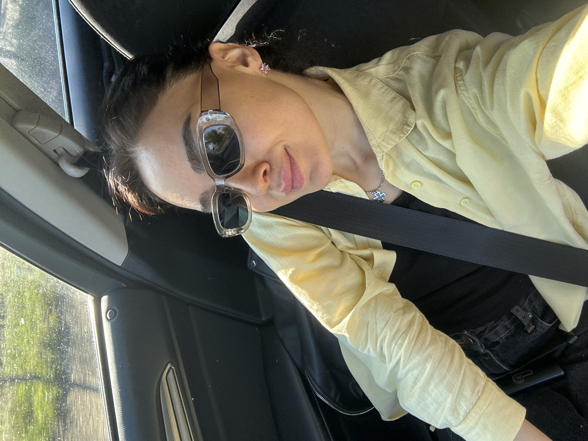 A person wearing sunglasses and a yellow shirt sits in a car. The person is wearing a seatbelt. A window is visible in the background, possibly indicating the location is in transit within the city of Tbilisi, Georgia, or elsewhere.