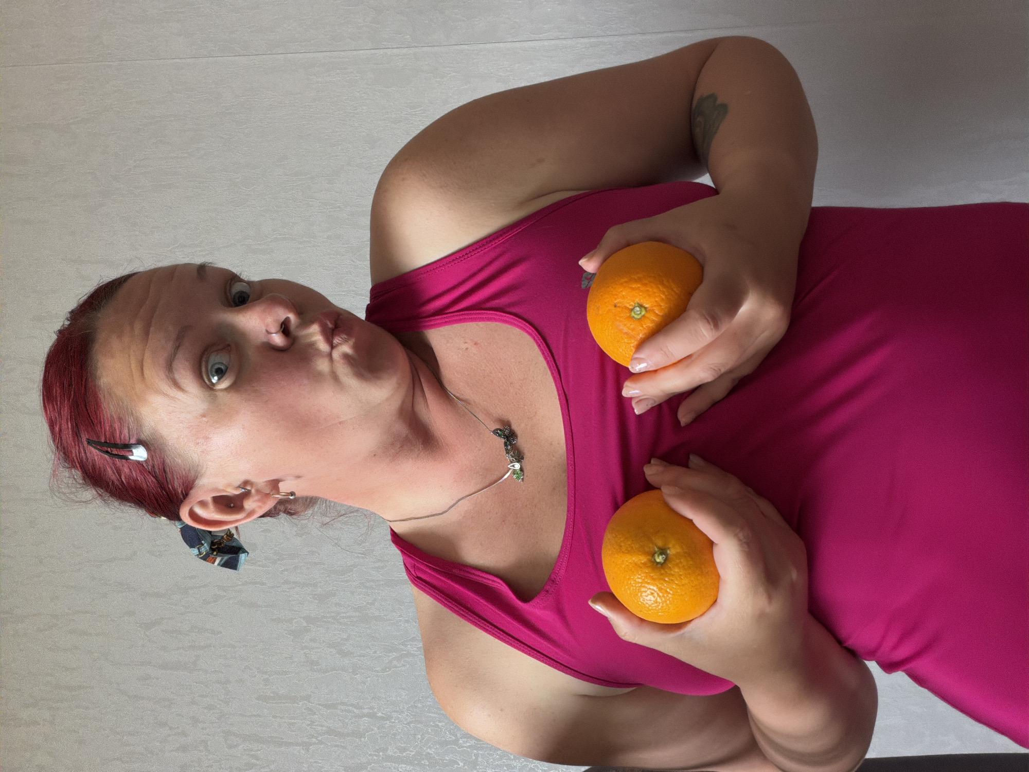 A woman with red hair and fair skin is posing with two oranges in front of a light-colored wall. She is wearing a pink tank top and has a silver necklace with a decorative pendant. She is holding one orange in each hand, positioned near her chest. Her facial expression is exaggerated, with pursed lips and wide eyes. There is a tattoo on her right upper arm. Her left ear has multiple piercings. There is a silver hair clip in her hair. The photo was taken at an angle. The location is Chaikovsky, Russia.