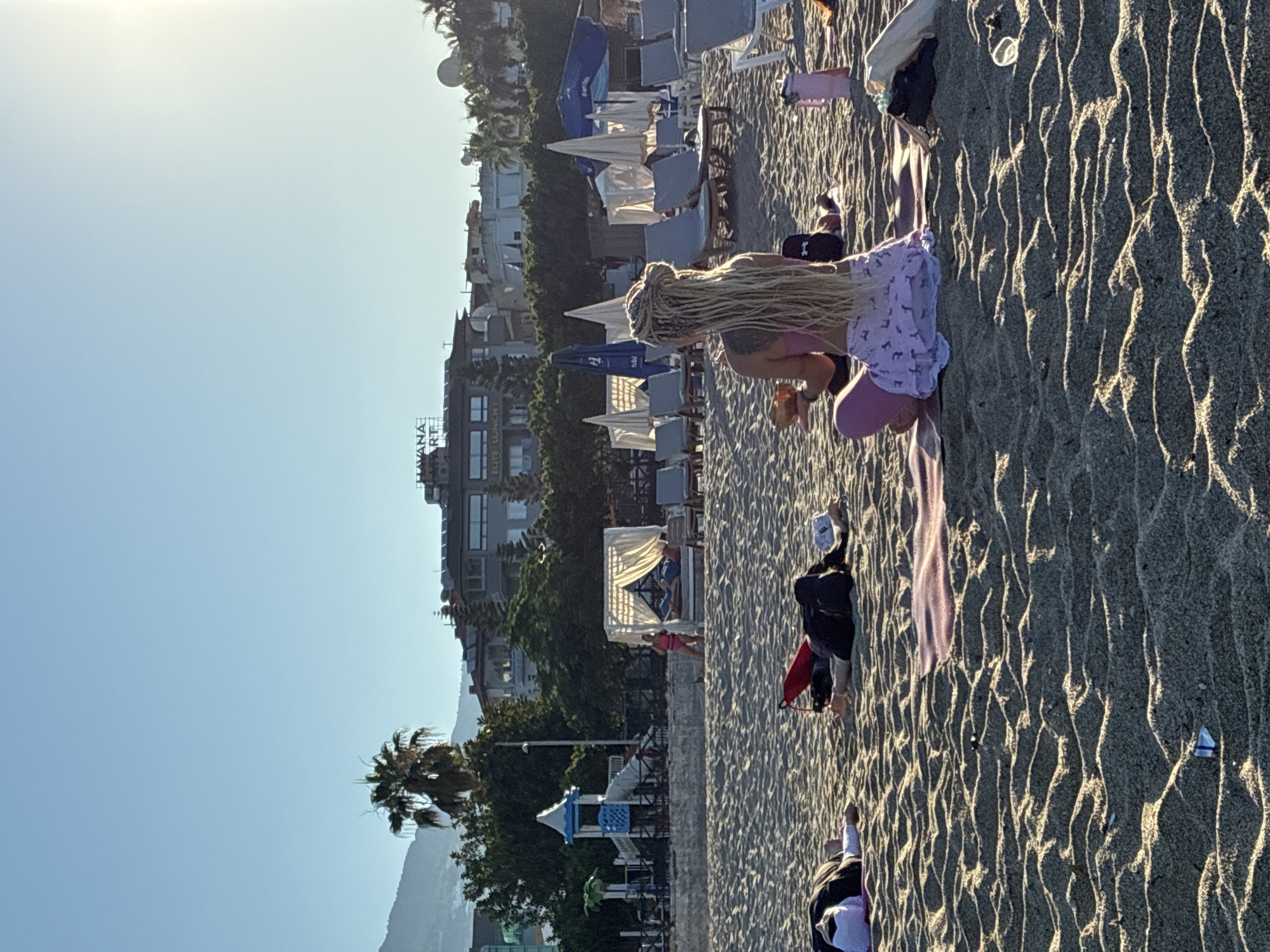 A group of people are on a sandy beach in Alanya, Turkey, with the sea and buildings in the background. In the foreground, a woman with blonde dreadlocks is sitting cross-legged on a yoga mat, holding a coconut and wearing a pink top and a light purple skirt. To her left, two people are lying on yoga mats, appearing to be resting or meditating. Further back, there are lounge chairs and umbrellas, suggesting a resort area. In the distance, there is a playground with a blue and white structure, and behind that, buildings are visible, including a hotel with a sign that reads "HAVANA RESORT." Palm trees are scattered throughout the scene. The sky is clear and blue, and the sun is shining.