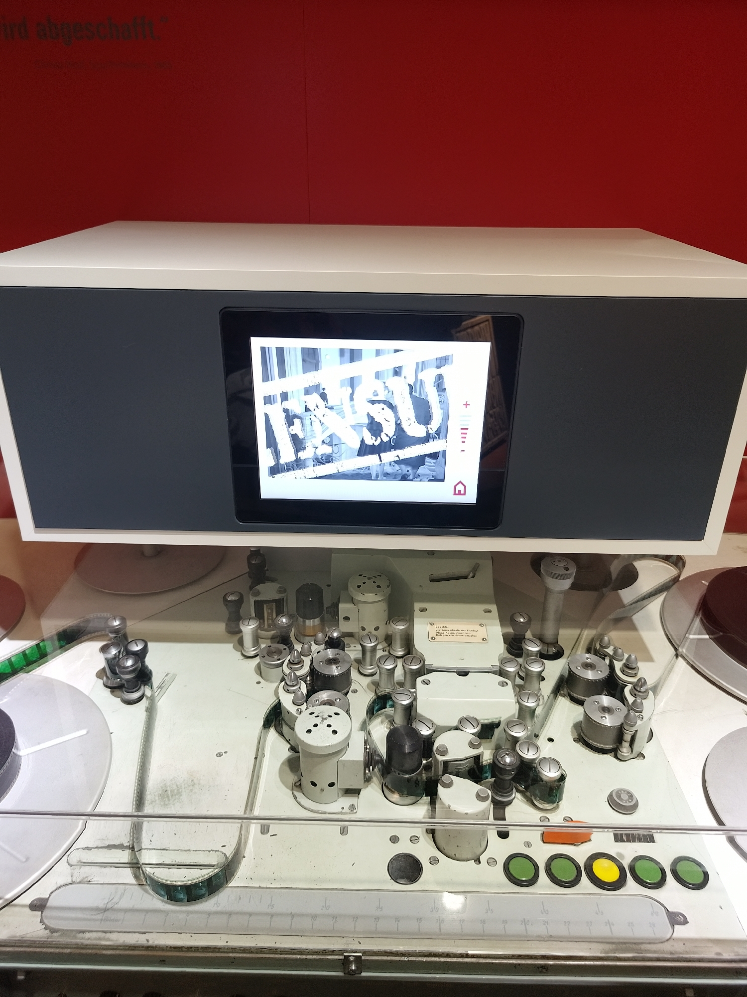 A film editing machine sits on display. The machine is white with various metal components including reels, spindles, and buttons. A screen shows a black and white image. The background is a solid red wall. The location is Unknown, Unknown.