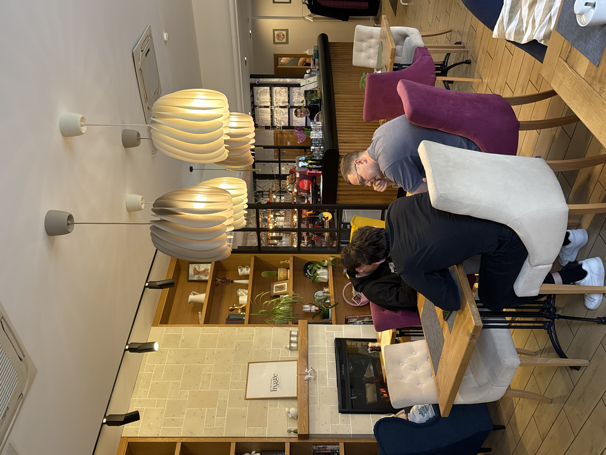 Inside a restaurant, two people sit at a wooden table with purple and white chairs. One person is wearing a gray shirt and glasses, the other a black jacket. They are indoors, near a fireplace and shelves displaying decorative items. The restaurant's interior includes distinctive light fixtures and a view toward the bar area. The location is Saint Petersburg, Russia.