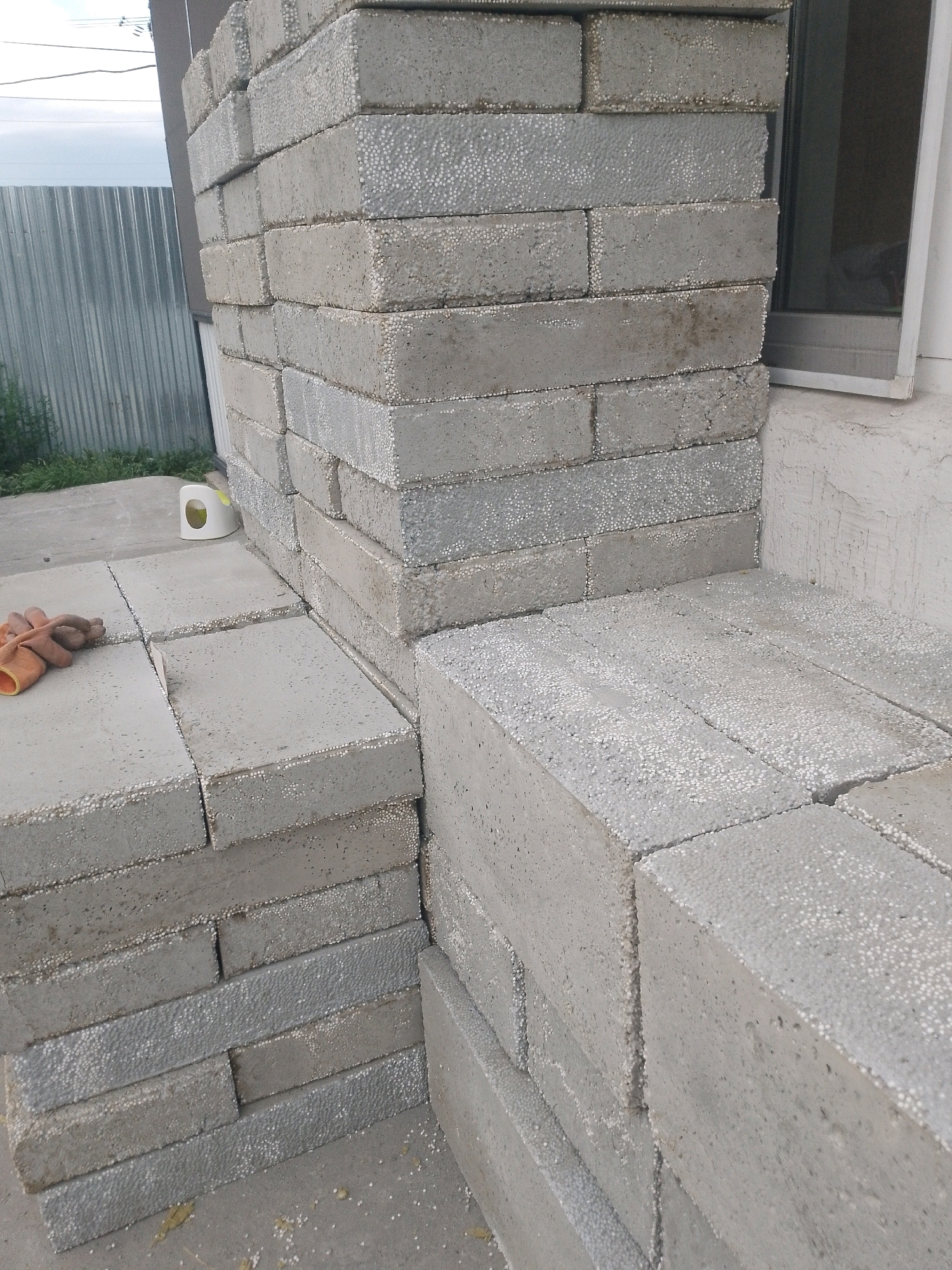 The image shows a stack of concrete blocks in an outdoor setting. The blocks appear to be made of aerated concrete or a similar lightweight material, characterized by visible small white pores. They are grey in color and are stacked in a corner formation, with some blocks forming a wall and others forming a horizontal surface.
In the foreground, on the left side of the image, there is a pair of work gloves lying on top of some of the blocks. Nearby, a small white object with a yellow opening is visible, possibly a dispenser or a small container.
In the background, to the left, a corrugated metal fence is visible, with some green vegetation growing near its base. Further in the background, a portion of a building with a window is present on the right side of the image. The sky is visible at the top, appearing overcast or cloudy. The ground beneath the blocks is a paved surface, likely concrete or asphalt. The overall scene suggests a construction or renovation project in progress.
The location is identified as Semey, Kazakhstan.