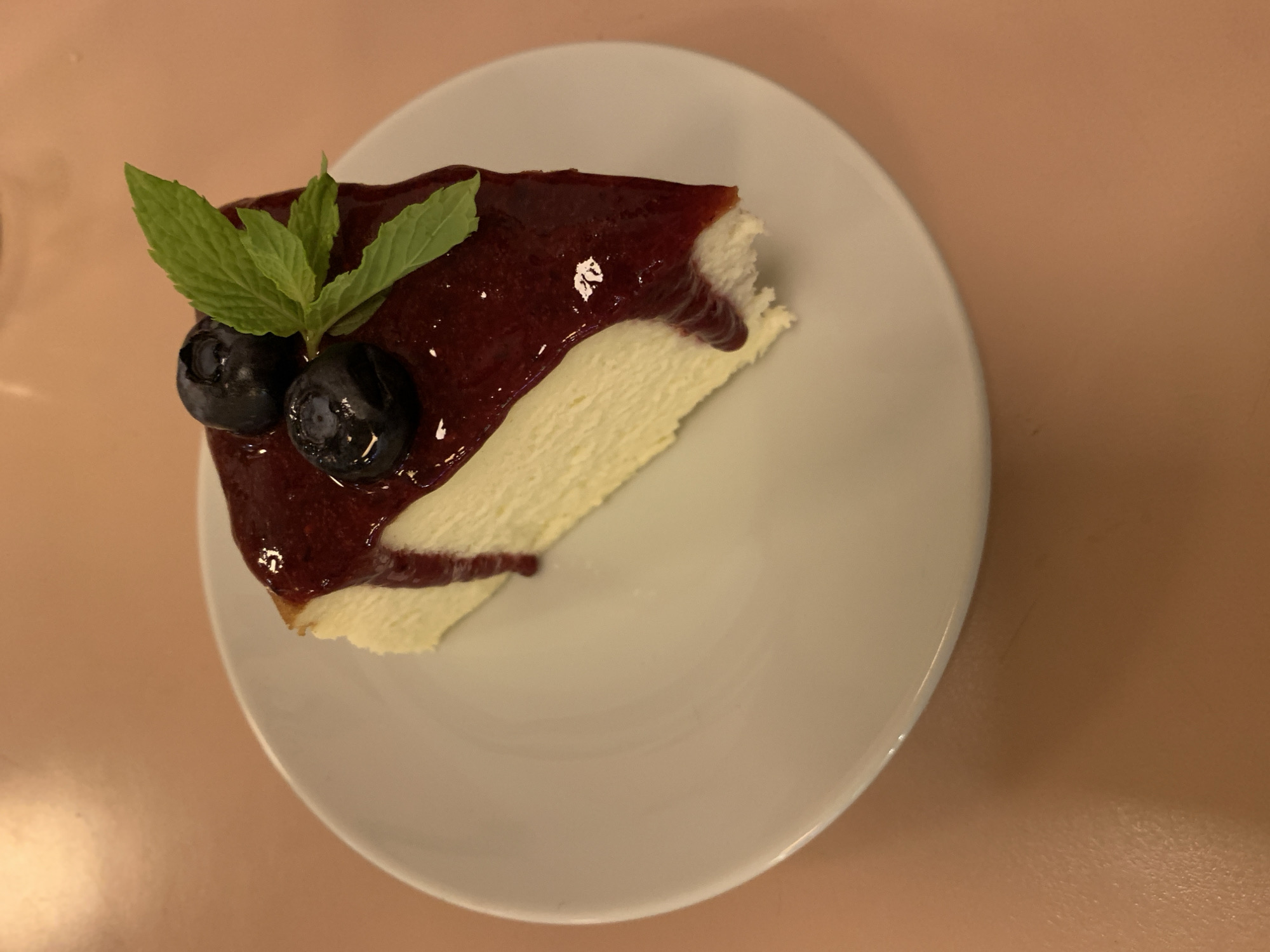 A slice of cheesecake with a dark red topping, two blueberries, and several green mint leaves sits on a white plate. The plate rests on a peach-colored surface. The scene appears to be indoors. No location in Buenos Aires, Argentina can be determined. No people, actions, or interactions are visible.