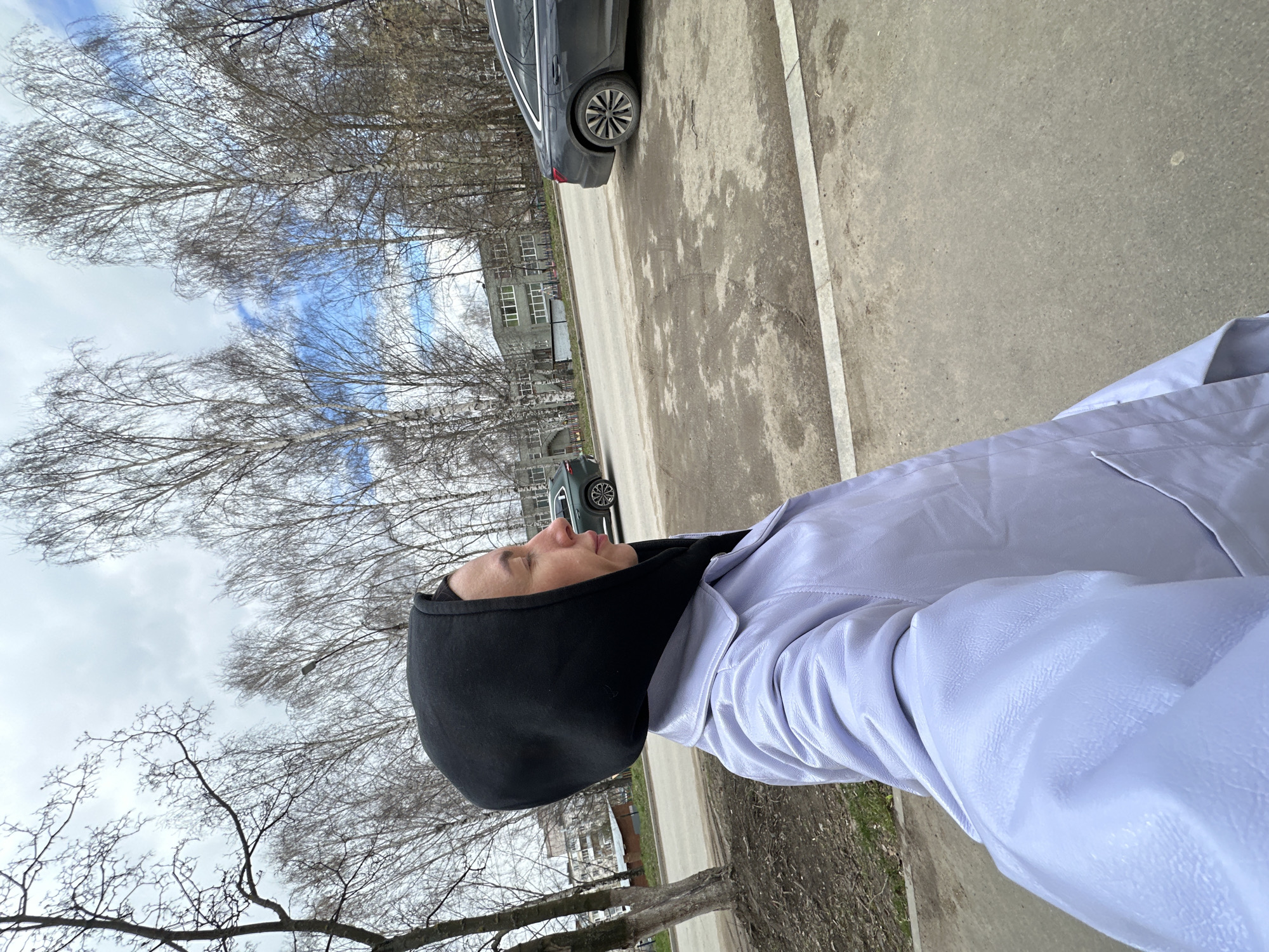 The image captures a daytime scene outdoors, likely in an urban or suburban environment in Votkinsk, Russia. The primary subject is a person, visible in profile from the chest up, positioned on the right side of the frame. They are wearing a black hood covering their head and a light purple jacket, possibly made of faux leather, with a textured surface. The person is looking slightly upwards and to the left, with a neutral expression.
The background is dominated by numerous deciduous trees with bare branches, suggesting either late autumn, winter, or early spring. Between the branches, patches of blue sky are visible, interspersed with white clouds. In the mid-ground, several multi-story buildings with multiple windows can be seen peeking through the trees. An asphalt road or paved path stretches across the mid-section and lower right of the image. A dark grey sedan is partially visible, parked on the side of this road, and another car is faintly seen further in the distance. The ground cover consists of dirt and some sparse vegetation near the trees, contrasting with the paved areas. There is no visible text in the image.