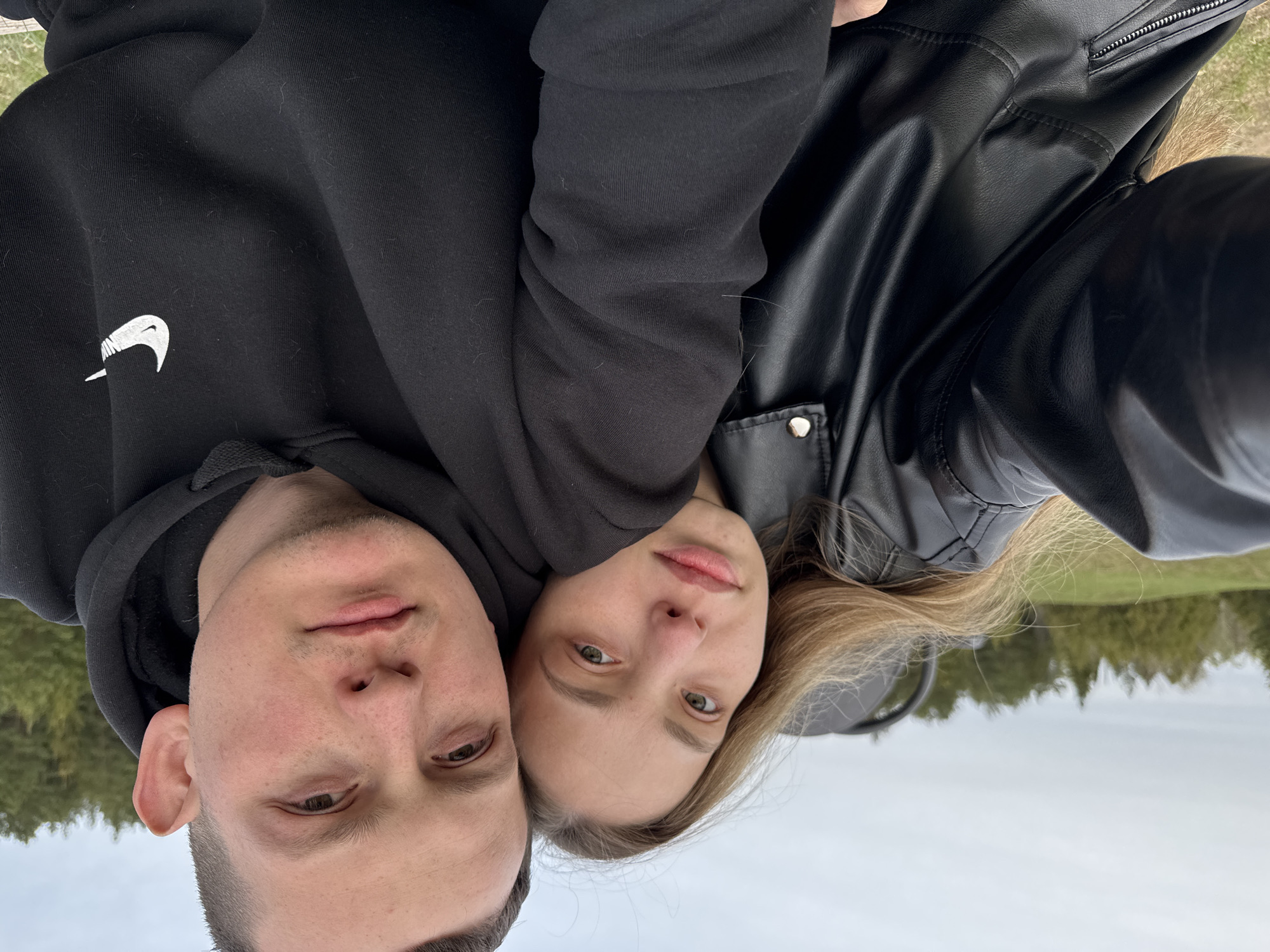 A photograph depicts a man and a woman closely positioned, possibly embracing. The man wears a black hooded sweatshirt with a visible Nike logo on the upper left side. The woman is dressed in a black leather jacket. The background reveals a landscape suggesting an outdoor location, potentially within the city of Ольховка, Russia, as evidenced by the presence of trees and sky. The overall impression is that of a casual, intimate moment captured in a selfie.