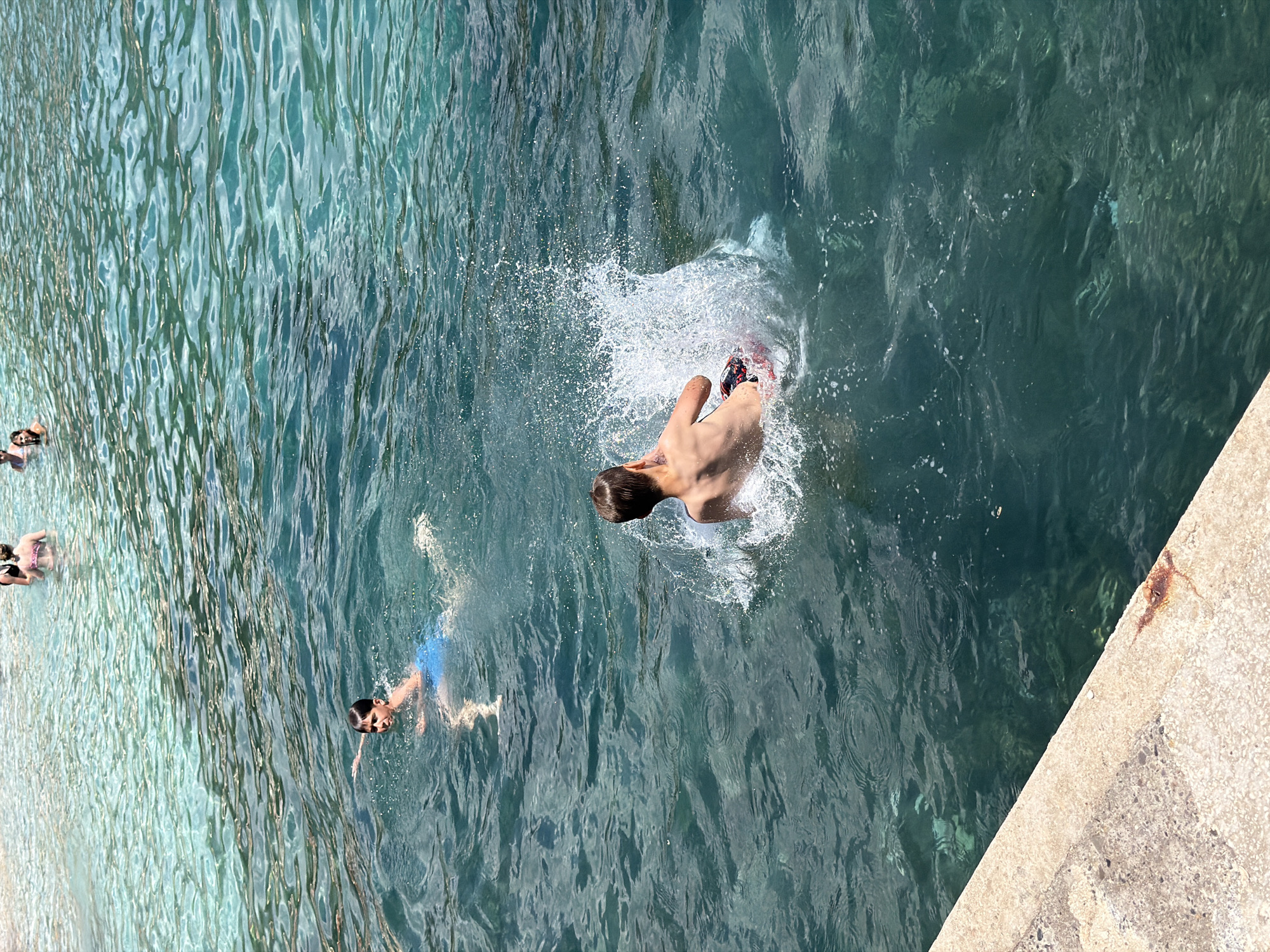 In Budva, Montenegro, two young boys are swimming in the clear turquoise waters of the Adriatic Sea. One boy, wearing blue swim trunks, swims towards the left of the frame, his arm extended as if reaching. The other boy, seen from the back, is in the process of jumping into the water, creating a large splash with his legs submerged. He is wearing patterned swim trunks. In the shallow, sunlit area further back, several other people are visible wading and swimming. The scene is set against a backdrop of the rocky coastline, with a concrete ledge in the lower left corner of the image.