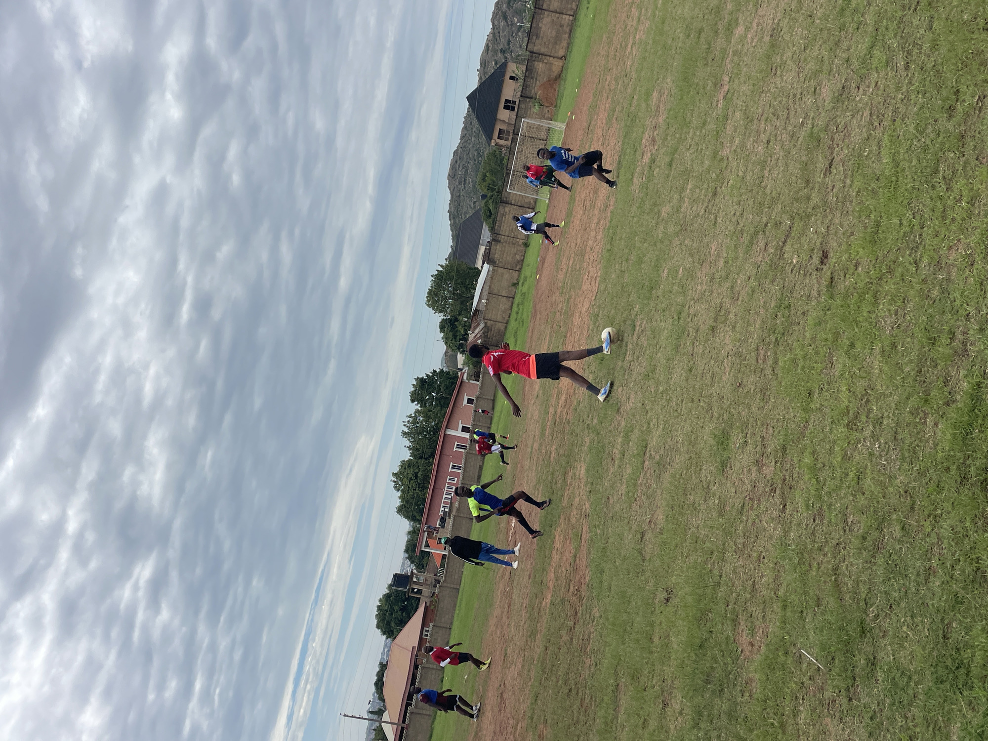The image depicts an outdoor football (soccer) match or practice session taking place on a natural grass field in Jos, Nigeria. The field shows areas of wear, with patches of reddish-brown soil interspersed with green grass.
The sky, which occupies the upper two-thirds of the frame, is largely overcast with extensive grey stratocumulus clouds. A narrow strip of lighter blue sky is visible near the horizon on the left side, indicating diffused daylight, likely late morning or early afternoon.
Eleven male individuals are visible on the field, most engaged in active movement consistent with playing football. Their attire consists primarily of athletic t-shirts, shorts, and athletic shoes in various colors including red, blue, yellow, black, and white.
In the left foreground, a player wearing a red shirt and black shorts is positioned with a white football near his right foot, appearing to prepare for action. To his right, in the mid-ground, a player in a blue and yellow top is captured in mid-stride, running towards the right side of the frame. Another player, in a blue shirt with a white logo, is running towards the foreground from the right side of the field. Further back, a player in a blue and white long-sleeved top is running towards the left. An individual dressed in a blue tracksuit with white stripes and white shoes stands relatively still in the mid-ground, observing the activity.
In the right background, a white metal goalpost with a net is visible on the field. Several small orange cones are placed on the ground near the goalpost.
The background of the scene features a series of residential or commercial buildings with red or dark-colored pitched roofs. These structures are partially obscured by a perimeter wall constructed of light-colored concrete blocks. Beyond the buildings, distinct rocky hills rise into the distance, particularly prominent on the right side of the frame. Scattered trees and shrubs are present throughout the background landscape.
