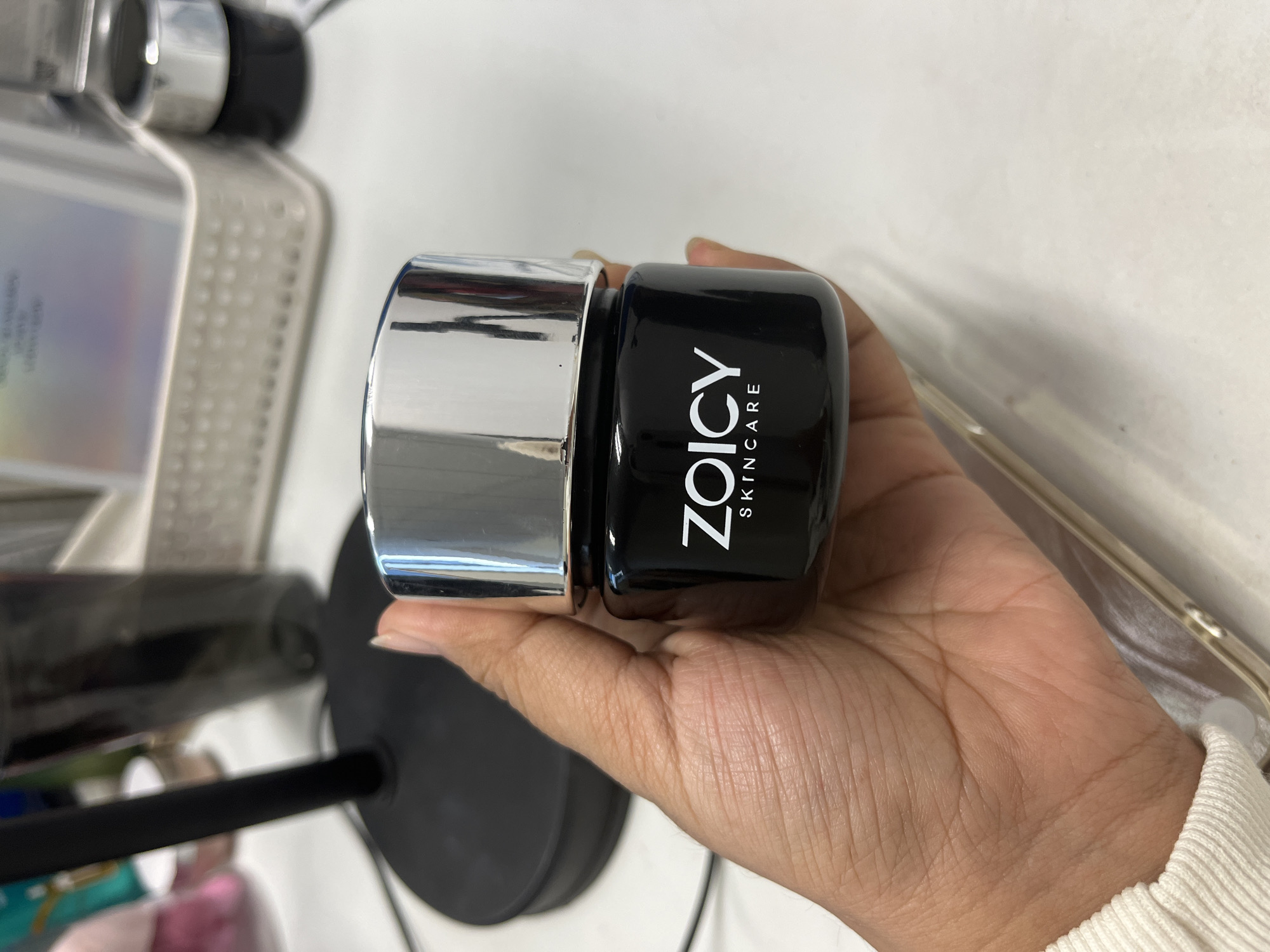 A close-up, eye-level shot shows a person's hand holding a small, black jar of ZOICY SKINCARE with a shiny silver lid. The jar is positioned in the center of the frame, with the hand grasping the bottom portion of the jar. The background is blurred, but appears to be a desk or countertop with office supplies and a black lamp base. The lighting is bright, casting shadows on the hand and jar. The location is specified as South Jakarta, Indonesia, but this is not visually evident from the image itself.