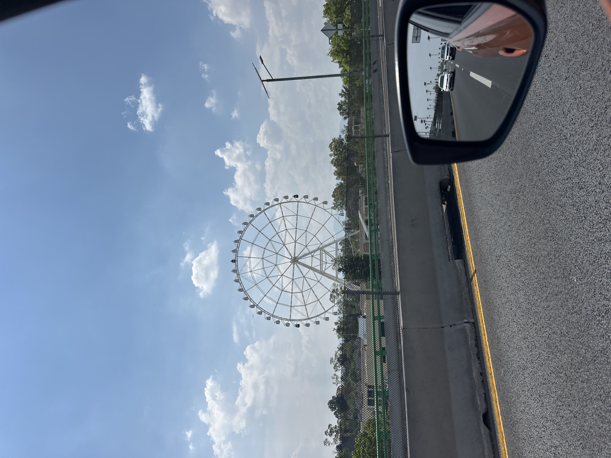 The image shows a scene from a vehicle traveling along a highway in Mexico City, Mexico. Visible through the side mirror is a view of the highway with multiple vehicles. A large Ferris wheel is on the left side, situated behind a barrier of fencing and trees. The sky above is partly cloudy, with a bright blue hue. The angle suggests the photo was taken from inside a moving car. The overall environment is urban, and the scene is captured during daylight.