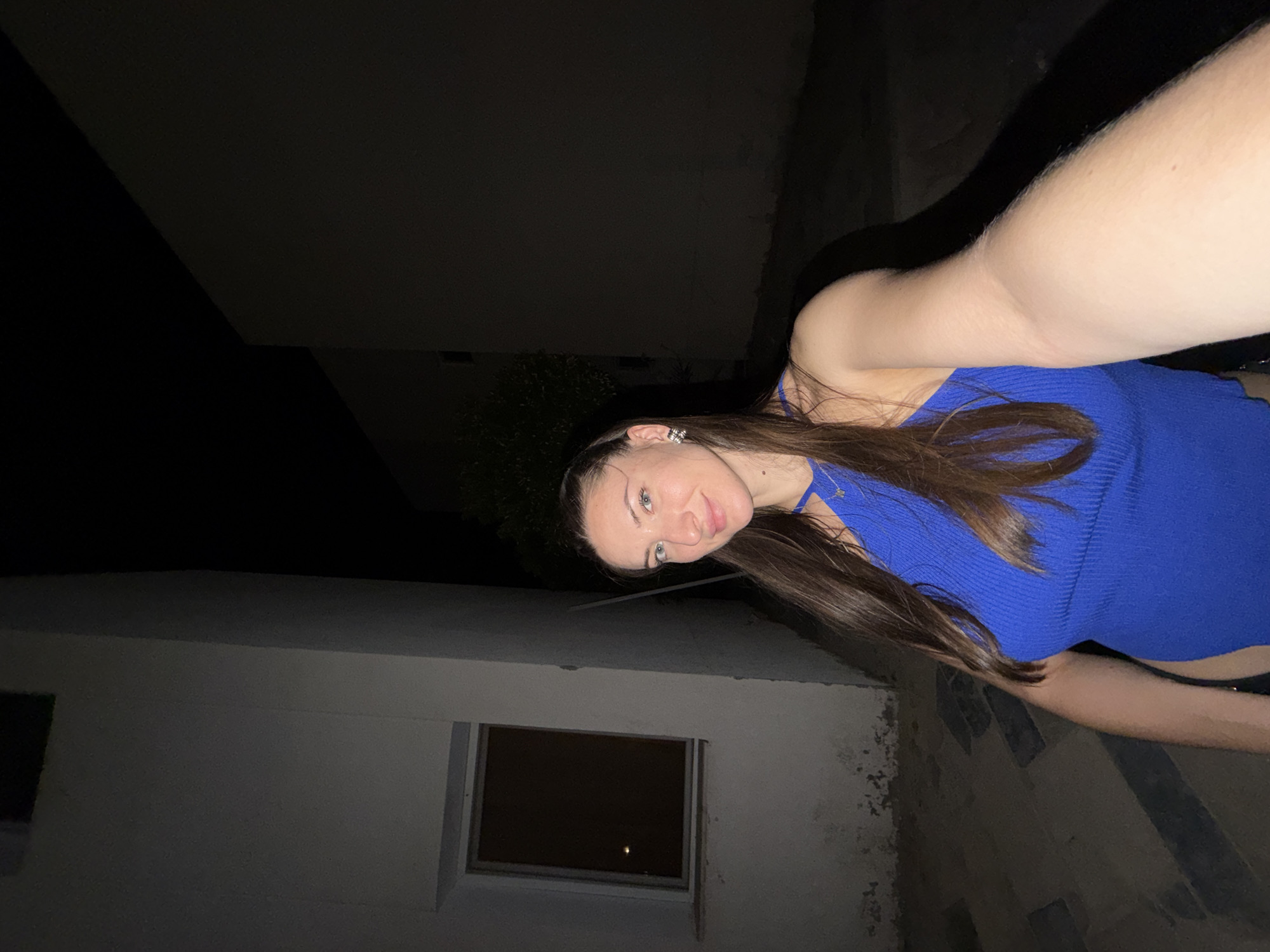 In the image, a woman with long, dark hair and blue eyes is taking a selfie at night. She is wearing a bright blue, ribbed top with a halter neck and a small gold necklace. She has silver earrings with black accents. The background appears to be an outdoor setting, possibly a courtyard or alleyway in Sharm El Sheikh, Egypt. To her left, there is a white building with a small, dark-paned window. The ground is paved with dark stones or tiles. The lighting suggests it is nighttime, with artificial light illuminating the woman and the immediate surroundings, while the background recedes into darkness.