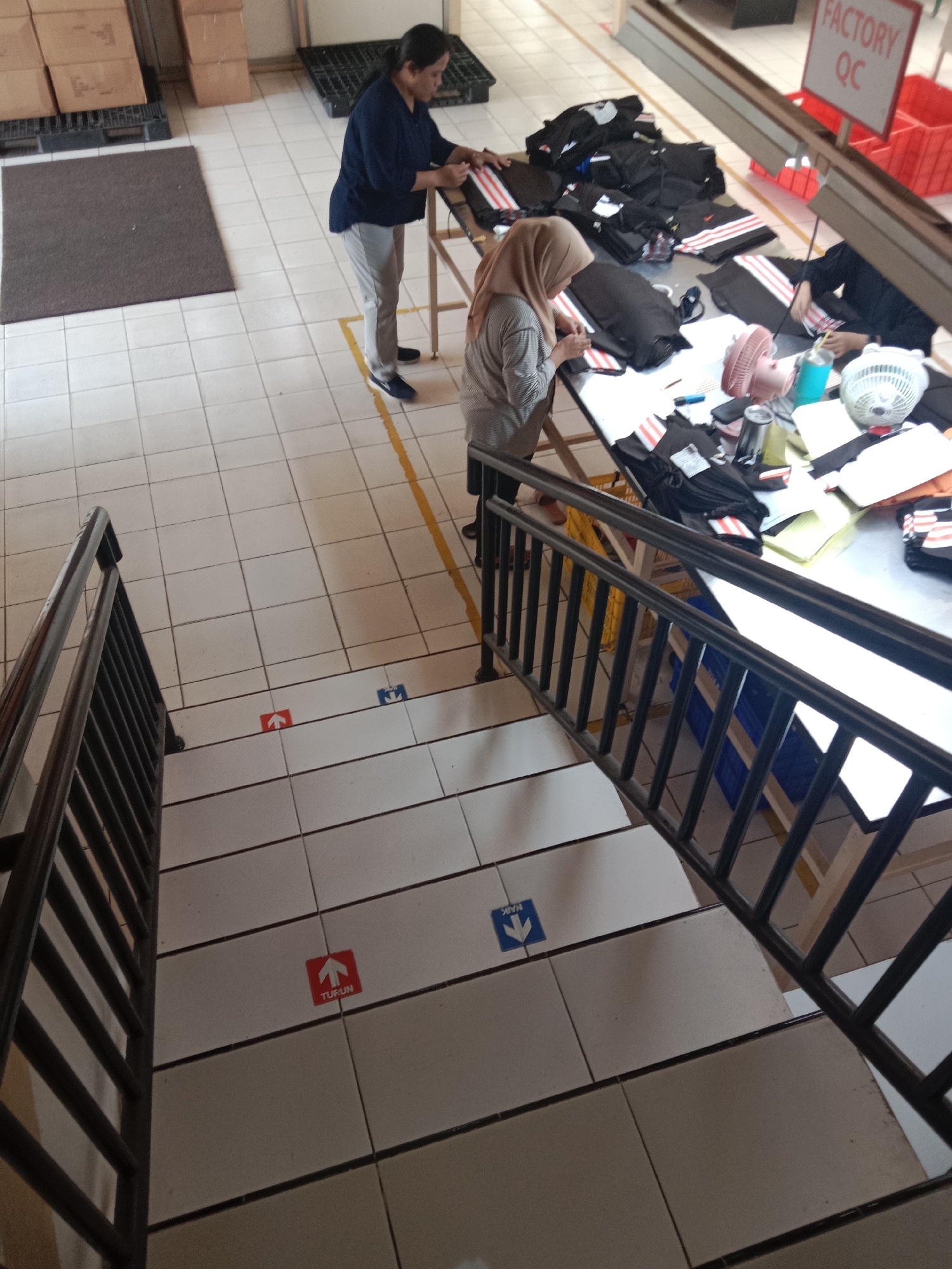 The image depicts a factory setting, likely in Semarang, Indonesia, with a focus on garment production and quality control. In the foreground, a black metal railing lines a tiled staircase, with directional floor stickers indicating "TURUN" (down) with a red arrow and "NAIK" (up) with a blue arrow.
Further up, a group of women are engaged in work at tables laden with black garments, some featuring white and red striped accents, possibly athletic wear. One woman, wearing a headscarf and a grey top, is seated and appears to be inspecting or assembling a garment. Another woman, in a dark blue top and light grey pants, stands at a table, also working on the garments. A third individual, partially visible, is also present at a workstation.
The environment includes tiled flooring, stacked boxes in the background, and various industrial items such as fans and containers. A sign labeled "FACTORY QC" is visible, reinforcing the context of quality control within the factory. The overall scene suggests an active production line environment.