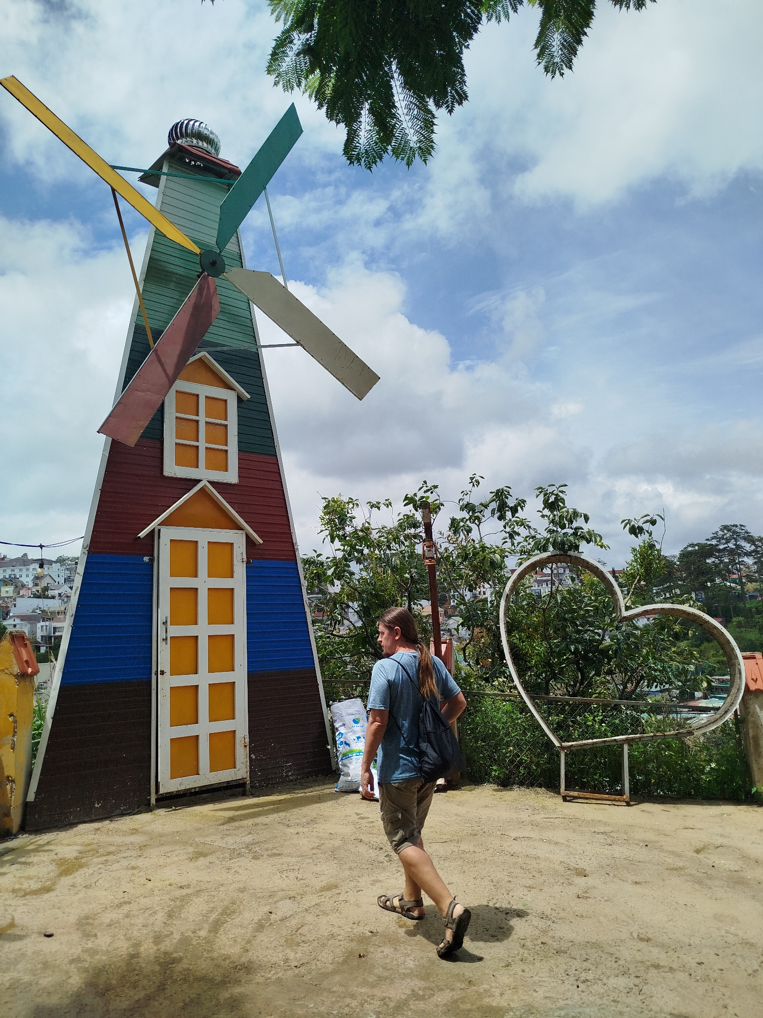 A person with long brown hair and a blue t-shirt is walking away from the camera, carrying a black backpack. They are wearing khaki shorts and sandals. The person is walking on a light brown, dusty path in front of a colorful, conical-shaped structure that resembles a windmill. The windmill has red, blue, dark brown, and green sections, with yellow doors and windows. Behind the windmill, a lush green hillside is visible with buildings scattered across it, indicative of the city of Da Lat, Vietnam. To the right of the windmill, a large white heart-shaped metal structure is present, partially obscured by green foliage. The sky above is a mix of blue and white clouds, with the fronds of a green tree hanging from the top of the frame.