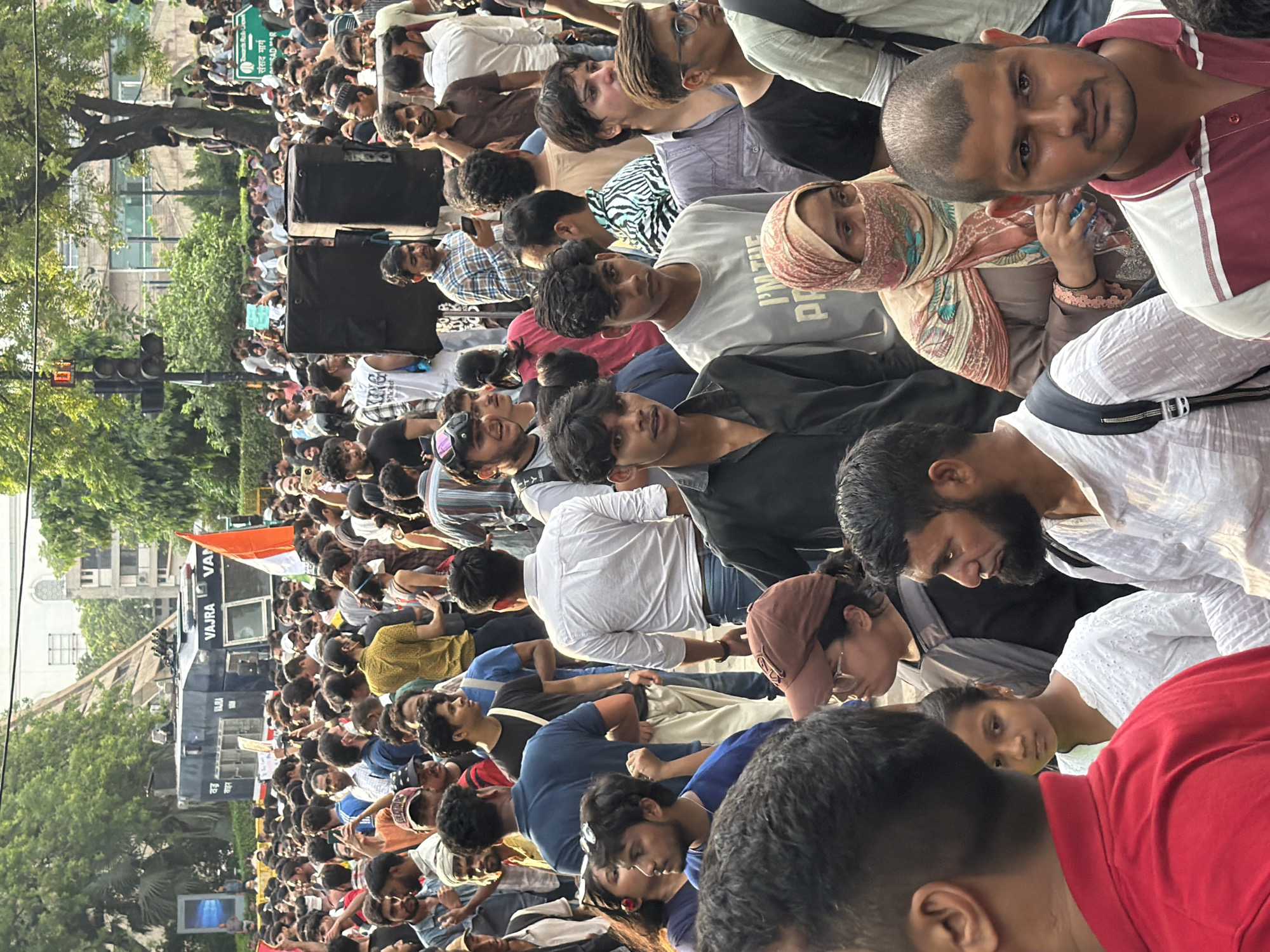 A large crowd of people, predominantly young men, are gathered outdoors in what appears to be a city street in New Delhi, India. The scene is busy and somewhat chaotic, with many individuals looking in various directions, some holding cell phones, suggesting they might be recording or communicating. In the background, a large, dark blue vehicle with the word "VAJRA" written on its side is visible, along with an Indian flag. There are also traffic lights and trees, indicating an urban setting. The people are dressed in casual clothing, a mix of t-shirts, shirts, and headscarves. Some individuals are looking directly at the camera, while others are engaged in conversation or observation. The overall atmosphere suggests a public gathering, possibly a protest or rally, given the presence of the vehicle and flag. The lighting indicates it is daytime.