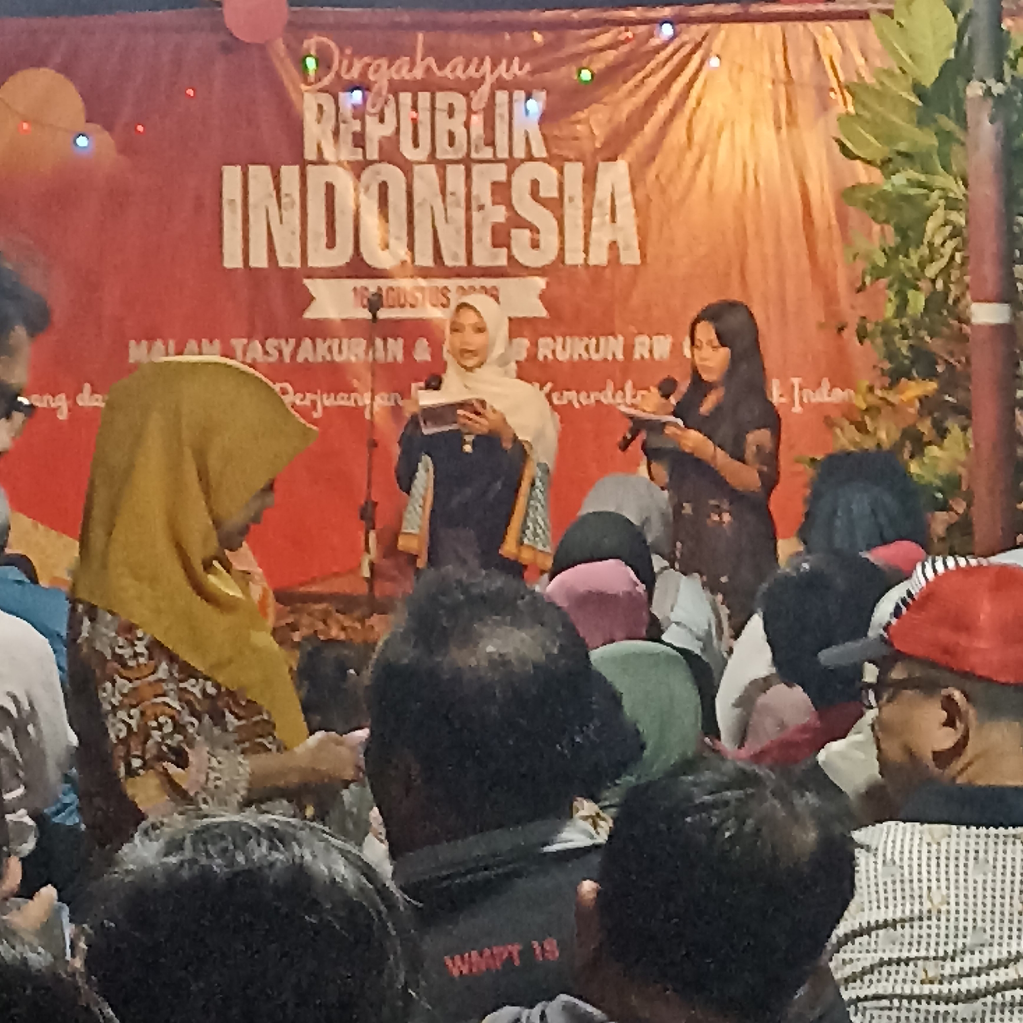 The image captures an outdoor event, likely a community gathering or celebration, taking place in the city of Magelang, Indonesia. A large red banner serves as the backdrop, prominently featuring the text "DIRGAHAYU REPUBLIK INDONESIA" in bold white letters, indicating a celebration of Indonesia's independence. Below this, smaller text reads "17 AGUSTUS 2020", specifying the date of the event.
In the center of the scene, two women are visible on a stage or elevated platform. The woman on the left, wearing a light-colored hijab and a dark dress with patterned sleeves, is holding a book and appears to be speaking or reading. To her right, another woman in a dark outfit is also holding a microphone and a document, suggesting she is also participating in a program.
A woman in a yellow hijab and a patterned dress is standing to the left of the stage, looking to her right. The foreground of the image is filled with the backs of a seated audience, their faces obscured, indicating their attention is directed towards the stage. The environment suggests a festive atmosphere, with decorative lights visible on the banner. The presence of lush green foliage on the right side of the frame hints at an outdoor setting, possibly a public space or community hall. The overall scene is one of a public celebration, likely related to Indonesian Independence Day, with community members gathered to participate.