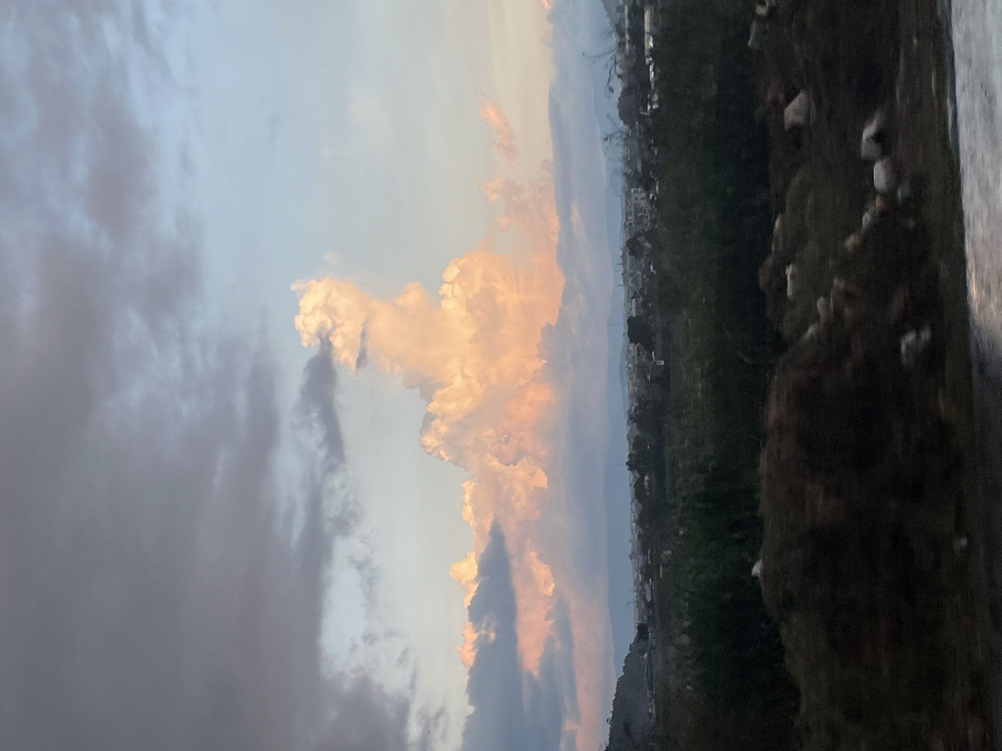 An eye-level, medium shot captures a landscape scene in Phi Nôm, Vietnam, featuring a dramatic sky with illuminated clouds. The sky is predominantly filled with large, fluffy clouds, some of which are tinged with pink and orange hues, suggesting either sunrise or sunset. Darker, grey clouds are present in the upper left portion of the frame, creating a contrast with the brighter clouds. Below the sky, a distant town or village is visible, with numerous buildings and structures interspersed with trees. The foreground consists of a slightly blurred area of dark earth and rocks, with hints of vegetation. The overall impression is a serene, natural landscape with a developing urban area in the background.