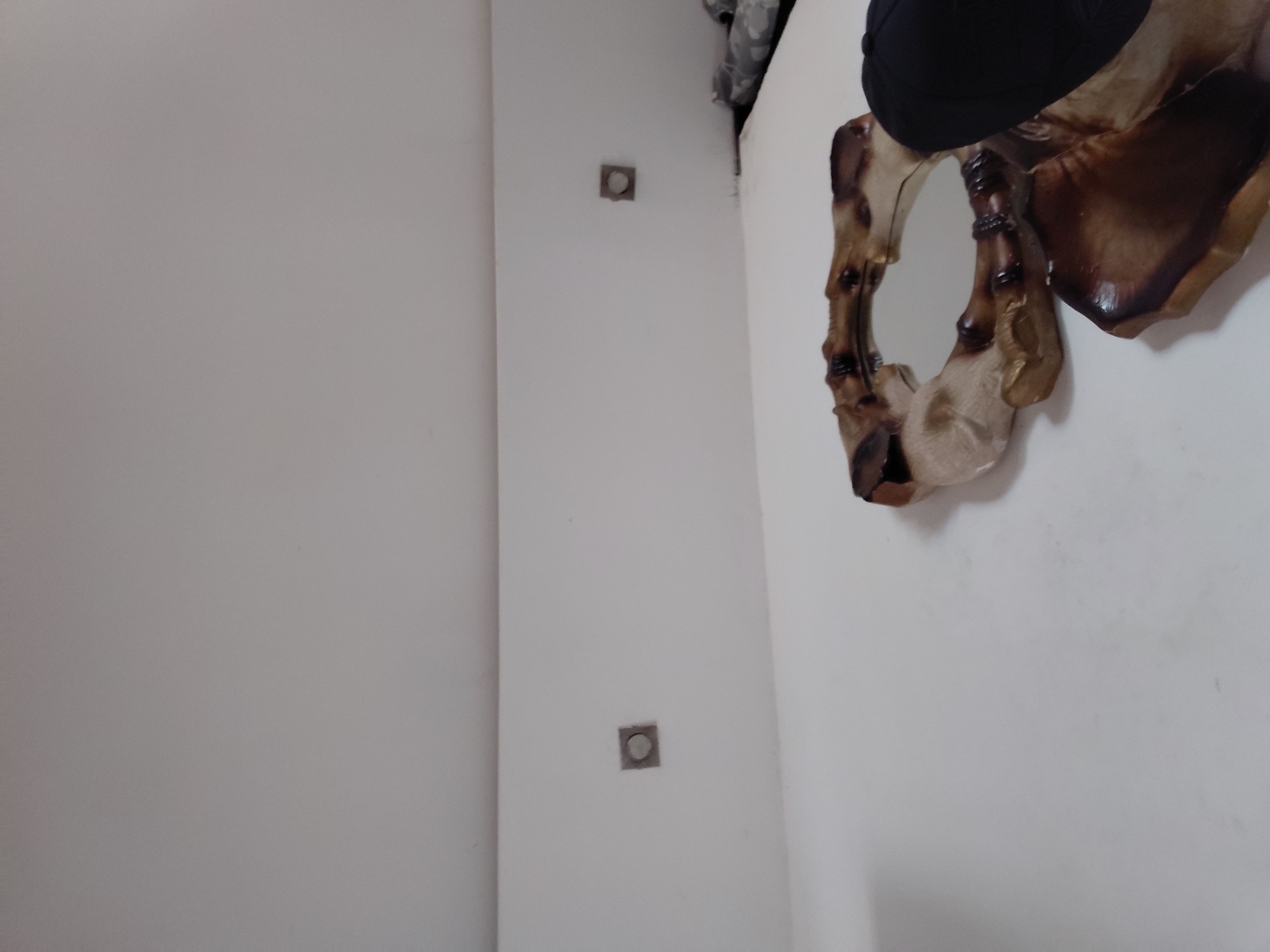 An indoor photograph, likely taken in Santa Ana, Venezuela, captures a static scene of a room with predominantly white or off-white walls. On the right side of the frame, a decorative mirror with a distinctive, organic-shaped frame is mounted on the wall. The mirror's frame appears to be crafted from or designed to mimic segmented wood or bone, featuring a mix of dark brown and lighter beige tones. A dark, possibly fabric item, resembling a cap or hat, is partially visible at the top edge of the frame, suggesting it might be resting on an unseen surface above the mirror.
To the left of the mirror, on a separate wall segment, two square-shaped wall fixtures are visible, vertically aligned. These fixtures are grey with a circular insert, appearing to be electrical outlets or possibly light switches. The overall lighting is even, typical of an indoor setting. No people are present in the image, and no specific activity or event is taking place. There is no visible text.