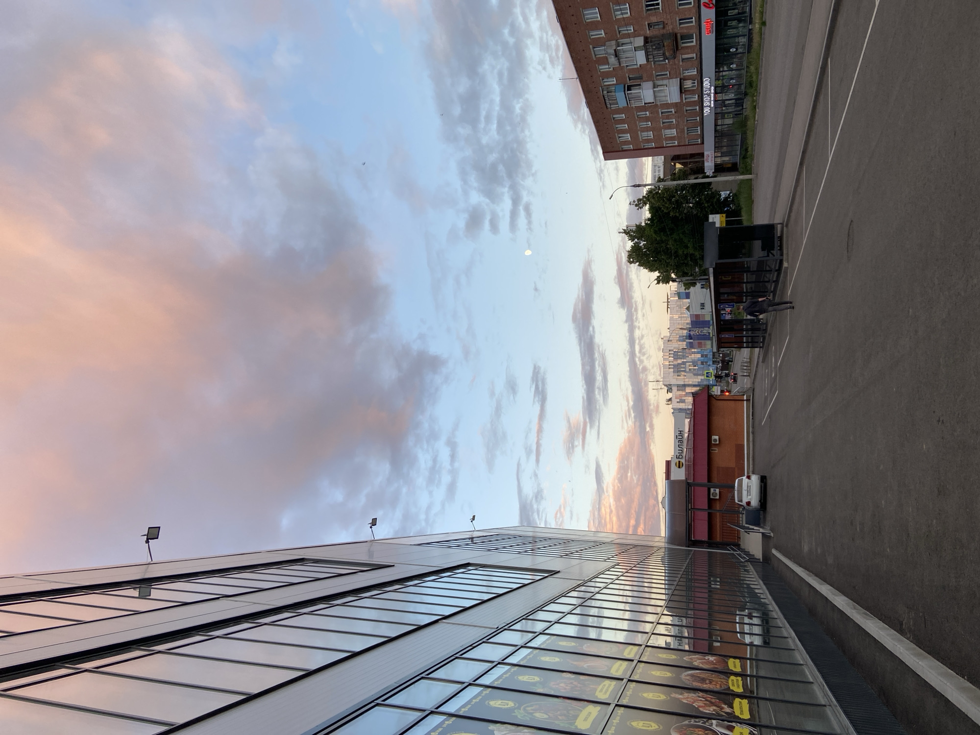 This is a vertical, outdoor, eye-level shot of a modern building with a glass facade, reflecting the sky and surrounding environment. The building is on the left side of the frame and extends into the distance. The sky above is partly cloudy, with soft hues of pink, orange, and blue, suggesting it's either dawn or dusk. A faint full moon is visible in the sky.
To the right of the modern building, there is an asphalt parking lot with white markings. In the distance, a multi-story brick building with many windows stands on the right side of the frame, partially obscured by a large green tree. Next to the brick building, there's a small black structure, possibly a bus stop or kiosk, with a person walking away from it towards the parking lot. Further down the parking lot, a white car is parked in front of a red brick building with signage.
The overall mood is peaceful and calm, with the soft lighting of either sunrise or sunset. The location appears to be an urban or commercial area, possibly in Votkinsk, Russia, given the context. There is some visible text on the buildings, but it is mostly illegible in the original image, except for "СУШИ" (SUSHISHOP) and "СУШИ СТУДИО" (SUSHI STUDIO) on the brick building, and what appears to be "БИЛАЙН" (BEELINE) on the red building.