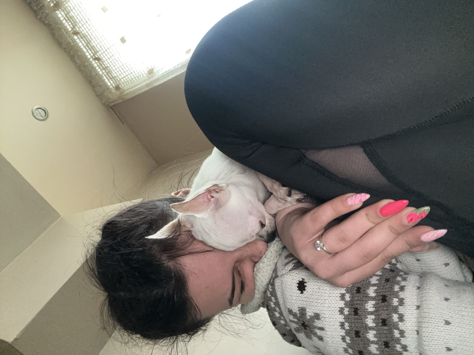 A woman is lying down with a white dog nestled on her face and shoulder. She is wearing a gray and white patterned sweater. The woman has dark hair pulled back and is wearing black leggings. Her fingernails are painted in a pink floral design. She has a ring on her finger. The location is Sliven, Bulgaria.