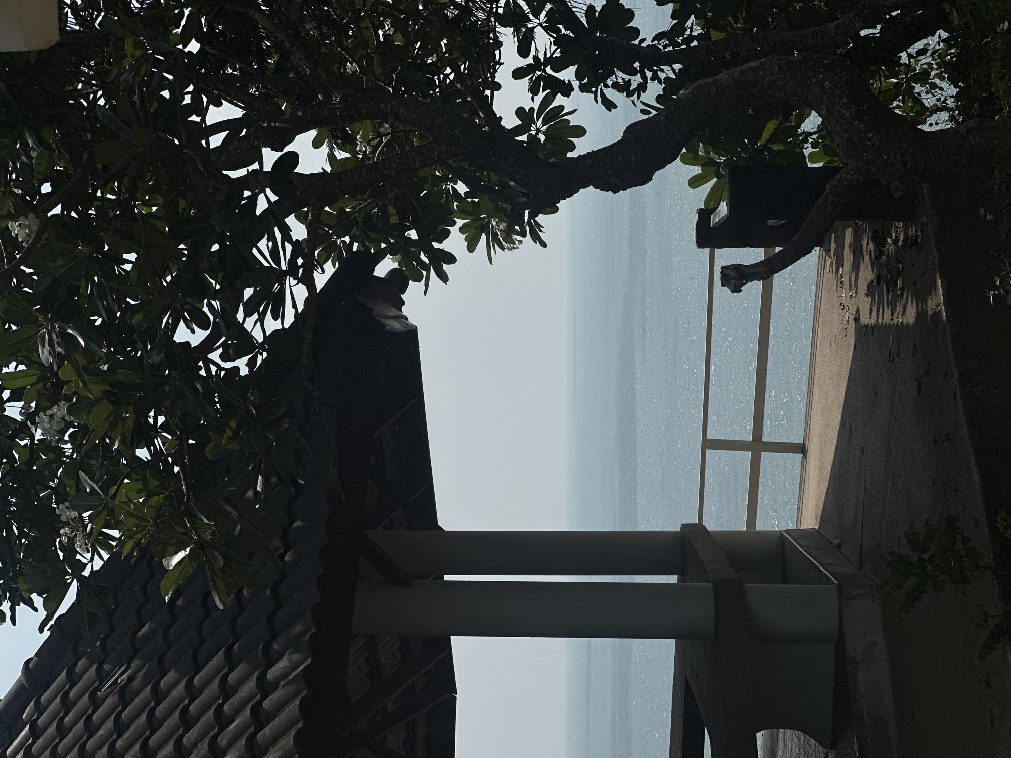 The image shows an outdoor scene, likely a balcony or veranda, overlooking a body of water. The view is partially framed by a tree with dense foliage at the top. The structure of the balcony consists of a dark tiled roof section in the lower left, white support beams, and a grey concrete railing. The body of water is slightly hazy. This image was taken in Ao Hin Khok, Thailand.