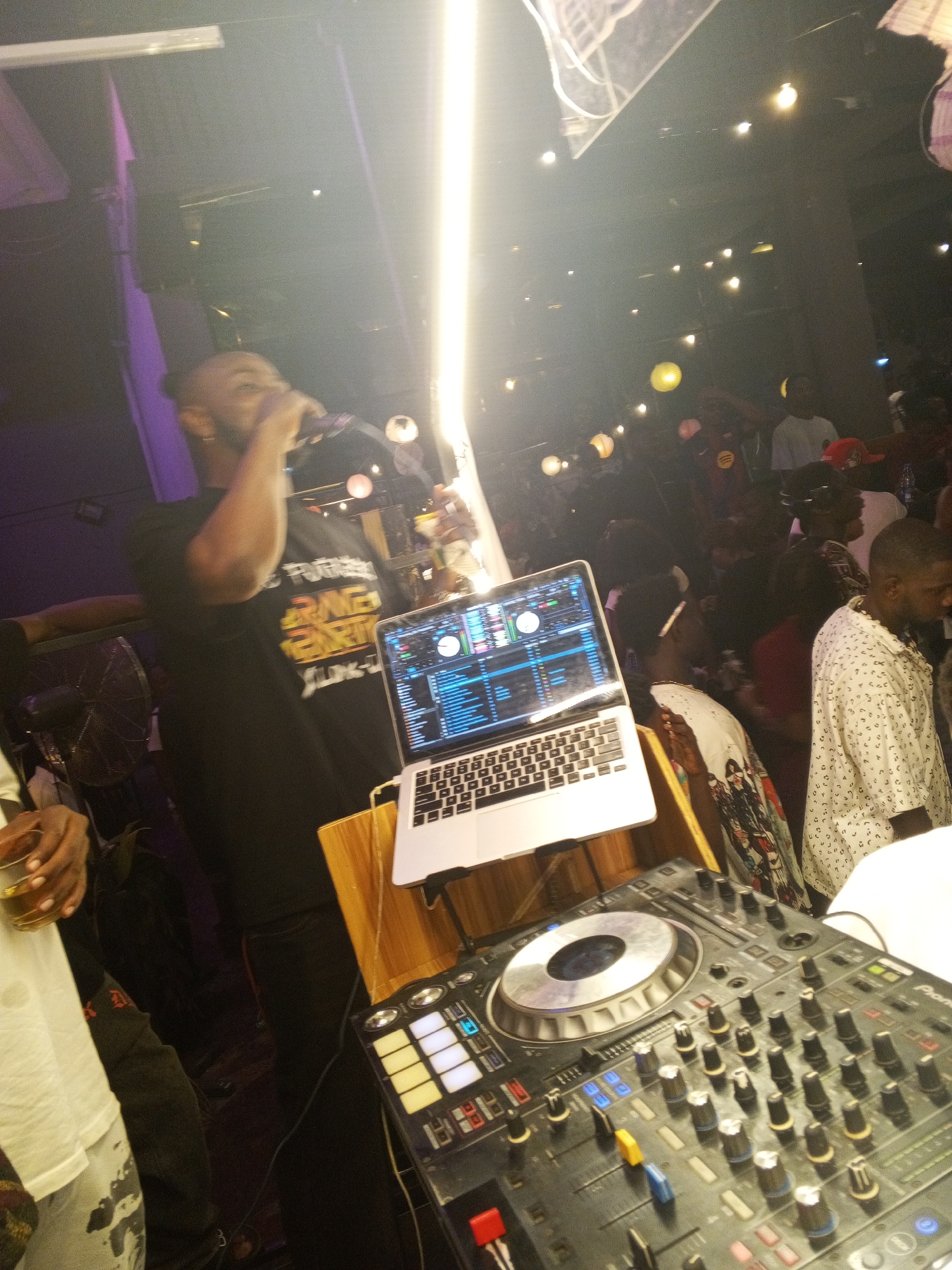The image shows a DJ performing in what appears to be a crowded nightclub. The DJ is a dark-skinned man wearing a black t-shirt with a colorful design, holding a microphone to his mouth. In front of him are DJ equipment, including a laptop computer displaying DJ software and a Pioneer DJ mixer. The lighting in the nightclub is dim with hanging lanterns and a bright light source above the DJ. Other people can be seen in the background, some of whom are looking at the DJ. The exact location in Lokoja, Nigeria cannot be determined based on the visual information.