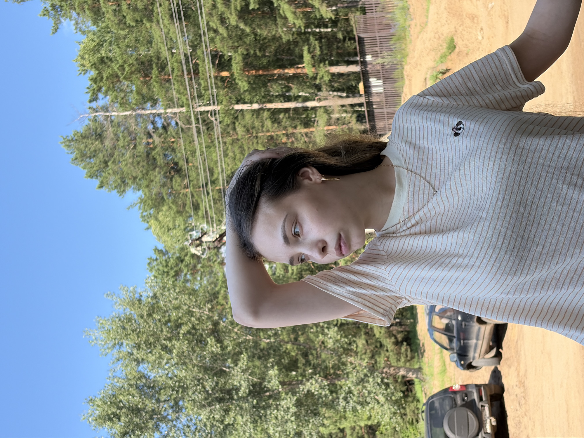 A young woman is standing in front of a wooded area and a dirt road. She is wearing a striped t-shirt and has her arm raised, holding her hair. Two cars are parked on the dirt road behind her, and a wooden fence is visible in the background. The sky is clear and blue. The image appears to be taken outdoors in a rural or semi-rural setting. It is not possible to determine the exact location within Noviy, Russia from the image alone.