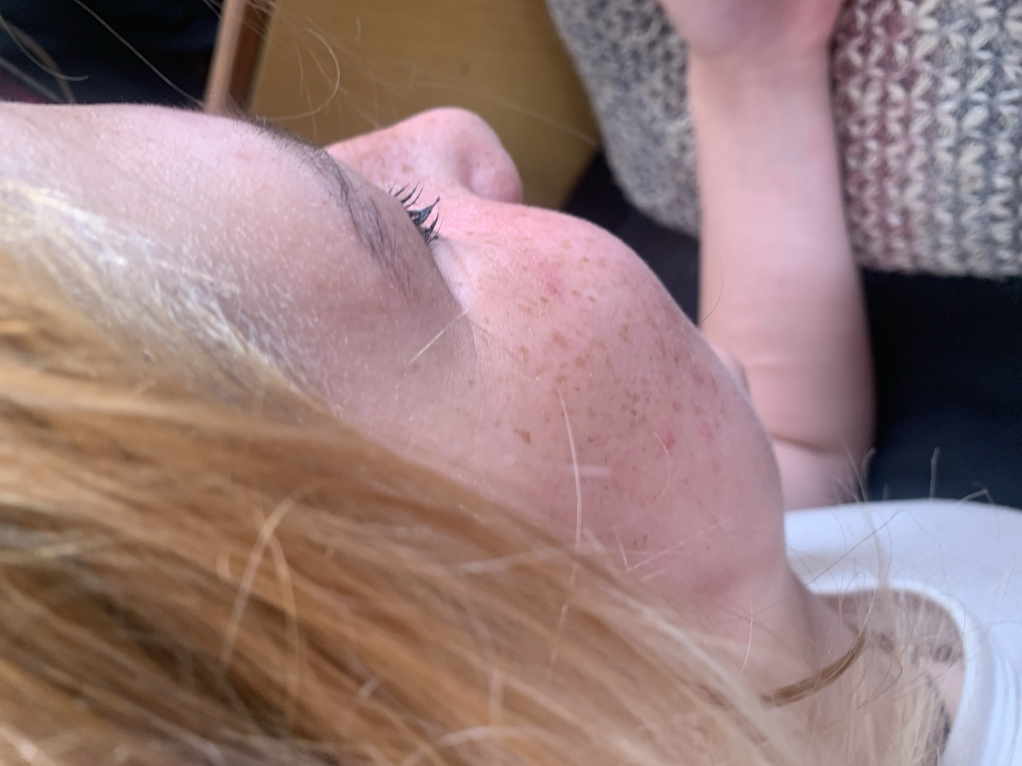 The image is a close-up, profile view of a person's face, specifically the right side, showing the cheek, nose, and eye. The person has fair skin with numerous freckles concentrated on the cheek and side of the nose. Their eyelashes are dark and appear to be made up. Strands of light blonde or reddish-blonde hair fall across the face and forehead. The background is out of focus, but appears to be an indoor setting with a warm-toned wooden surface and a textured fabric that could be a blanket or upholstery. There is no visible indication of the city of Zhmerynka, Ukraine.
