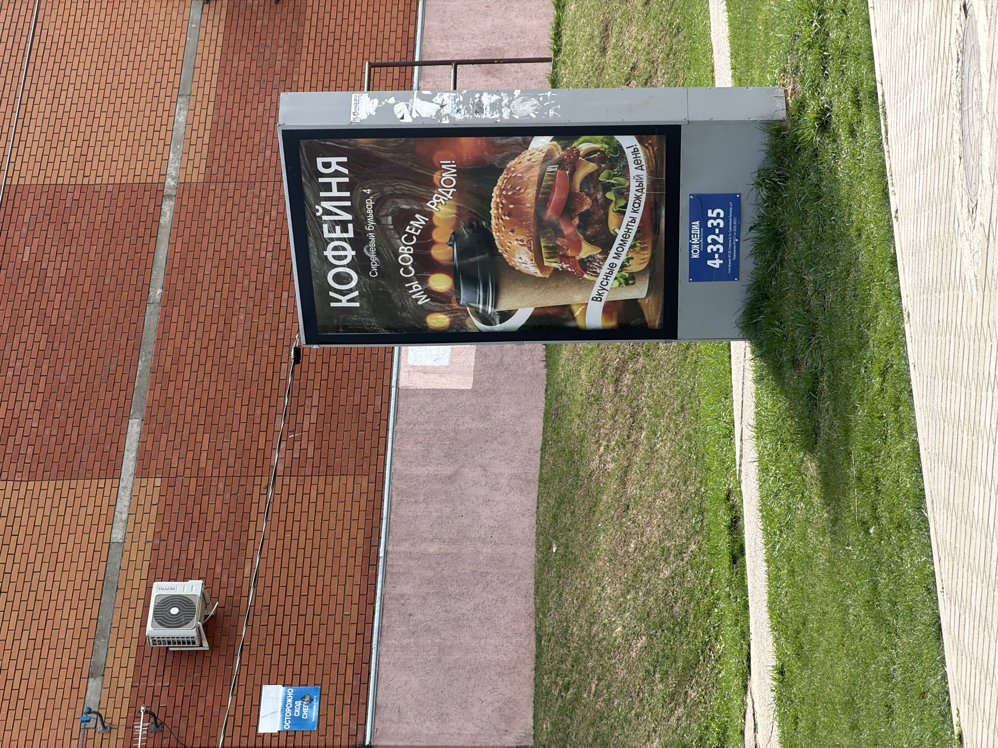 The image depicts an advertising billboard located outdoors, likely in a public space, possibly Chaikovsky, Russia. The billboard promotes a "Kofeynya" (coffee shop) located at 4 Sirenevyi Boulevard. The advertisement features a stylized image of a burger. Additional text states, "We are very close!" and "Delicious moments every day!" The billboard also displays the phone number 4-32-35 and mentions "KOK MEDIA." A portion of a brick building with an air conditioning unit is visible to the left. A caution sign that reads “ОСТОРОЖНО Сход СНЕГА” (Careful Snow Coming Down) is also visible on the wall. The billboard is positioned next to a grassy area. A walkway is visible on the right side of the image.