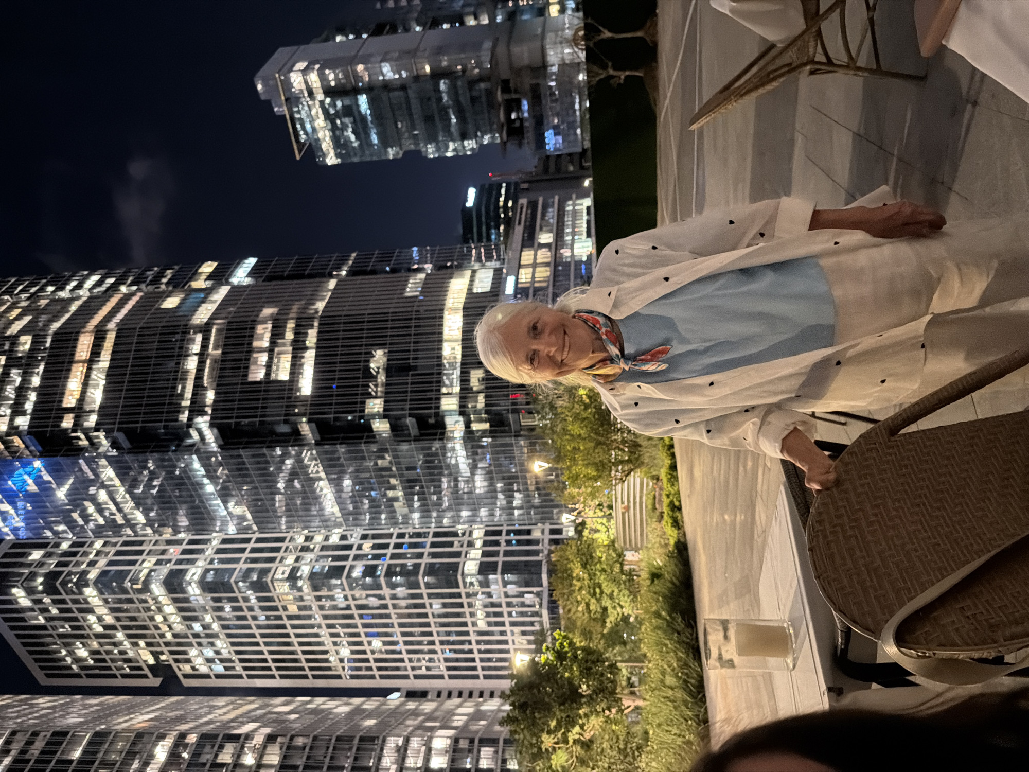 In Tel-Aviv, Israel, at night, a woman with white hair and a light blue shirt, white open jacket with black heart patterns, and light-colored pants smiles at the camera. She is standing on an outdoor patio with a wicker chair in the foreground. Behind her, modern skyscrapers with lit windows illuminate the night sky. In the middle ground, trees and greenery are visible, along with streetlights. A candle in a glass sits on a table to her left. The scene appears to be a relaxed social gathering in a well-lit urban setting.