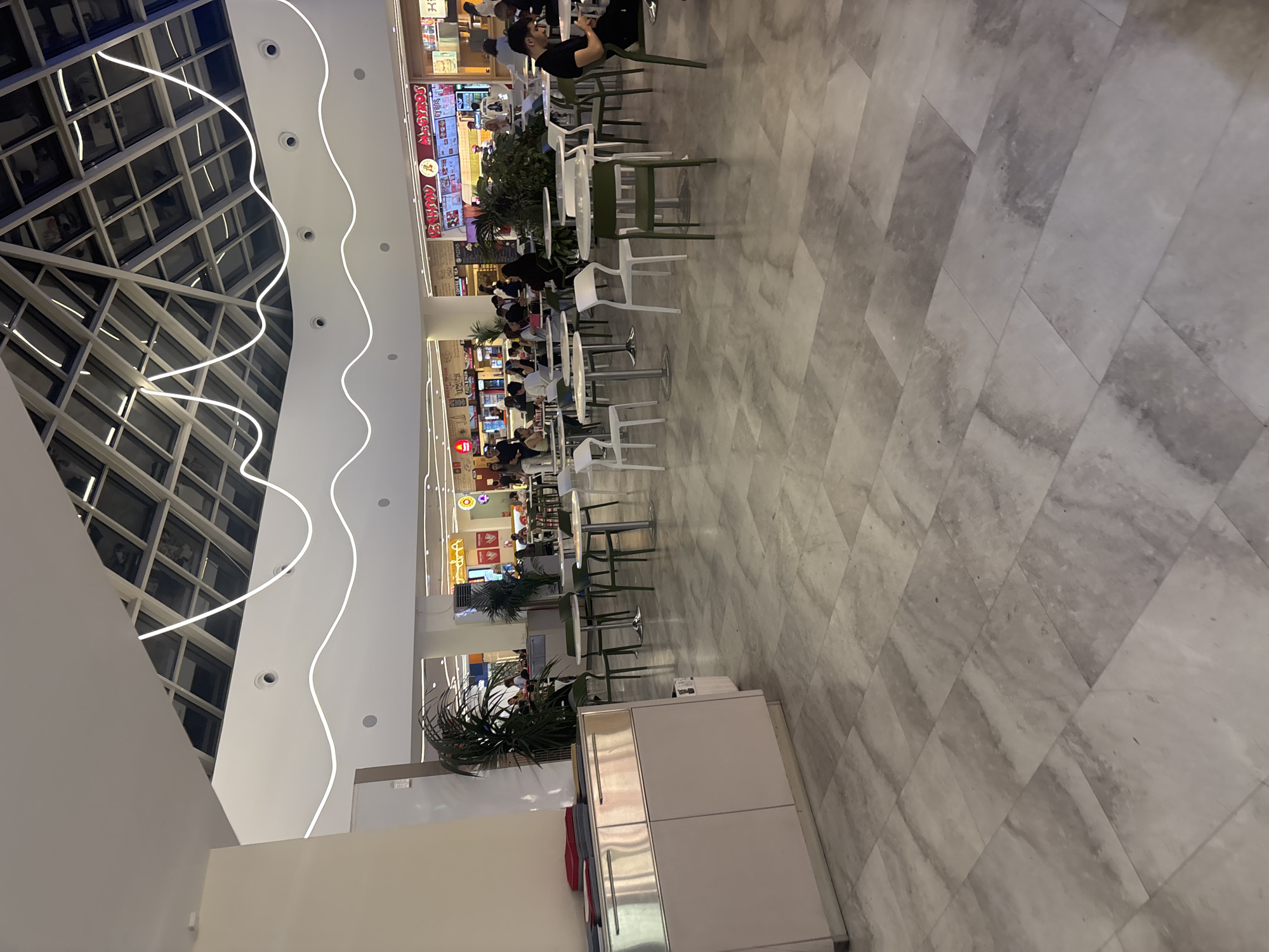 This image depicts a brightly lit, modern indoor food court or common area within a large commercial complex, likely a shopping mall, in Yerevan, Armenia. The scene is viewed from a slightly elevated perspective, looking down into the bustling area.
**Subjects:** Numerous people are present, primarily seated at tables, engaging in dining and conversation. They are casually dressed and appear relaxed. Some individuals are standing or walking between tables.
**Objects:** The area is furnished with a variety of modern tables and chairs. There are round and square tables, accompanied by both white and dark green chairs with simple, contemporary designs. Several large potted plants, resembling palm-like bushes, are strategically placed throughout the seating area, adding greenery. In the background, multiple food stalls or restaurant counters are visible, featuring illuminated signs and menus. In the foreground on the right, a long, low, light-colored service cabinet with reflective metallic elements is visible, with stacked red plastic baskets beside it.
**Scene and Setting:** The architecture is striking, with a high ceiling featuring a distinctive grid-like structure, possibly a skylight or an artistic ceiling installation, on the left. The ceiling also boasts decorative, wavy LED strip lighting that runs across its white surface, complemented by numerous recessed spotlights. The flooring consists of light-colored, possibly marble or tiled, material laid in a diagonal pattern. The overall ambiance is clean, spacious, and contemporary.
**Activity or Event:** The primary activity is people dining, socializing, and moving through a food court environment. It captures a typical moment in a public dining area during what appears to be daytime or early evening, given the bright interior lighting.
**Notable Details:** The architectural elements, particularly the ceiling design and wavy LED lights, are distinctive. The food court features multiple vendors.
**Visible Text:** Several signs for food establishments are visible in the background. One prominent sign clearly displays "MEGADZOS" in Latin script, with "SHAWARMA" also partially readable below it. Additionally, Armenian script is visible on multiple signs, confirming the location context of Yerevan, Armenia. Other signs display various graphics and menu items, though the text is largely unreadable due to distance and angle.