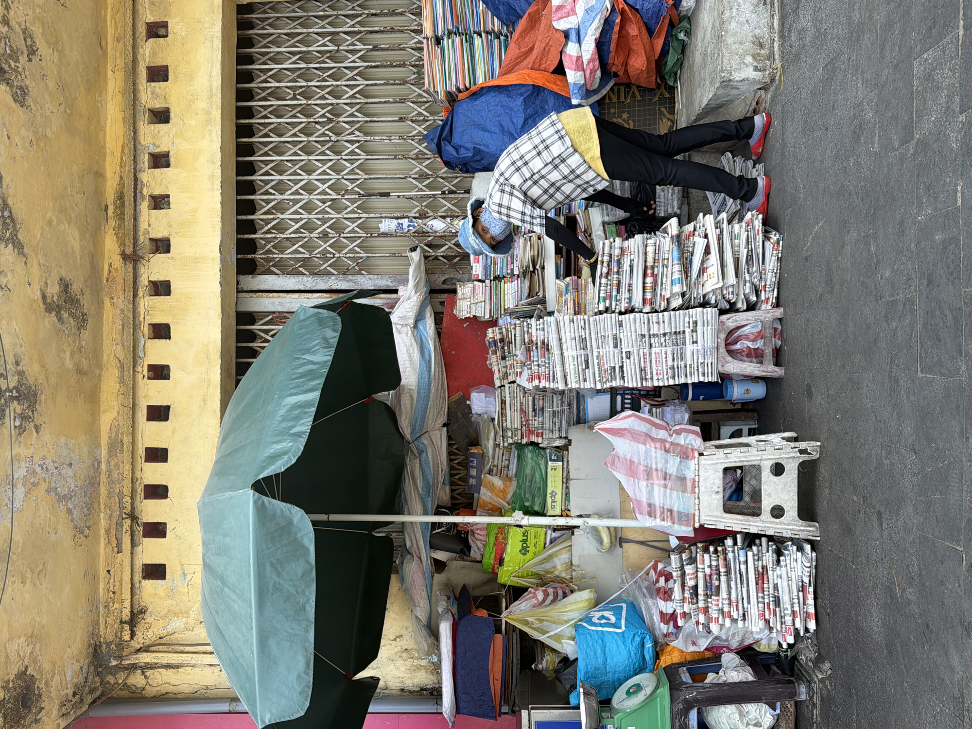 A ground-level, slightly downward-angled shot captures a vendor and their wares on a sidewalk in Đà Nẵng, Vietnam. The vendor, wearing black pants and red shoes, leans forward amidst stacks of newspapers and books. They wear a light blue hat and a plaid jacket with a yellow undershirt. A large, greenish-blue umbrella is positioned to the left, providing shade. Behind the vendor and their goods is a closed storefront, its metal shutter drawn down. The building is painted yellow. The sidewalk is gray. The scene suggests a street-side bookstore or newspaper stand.