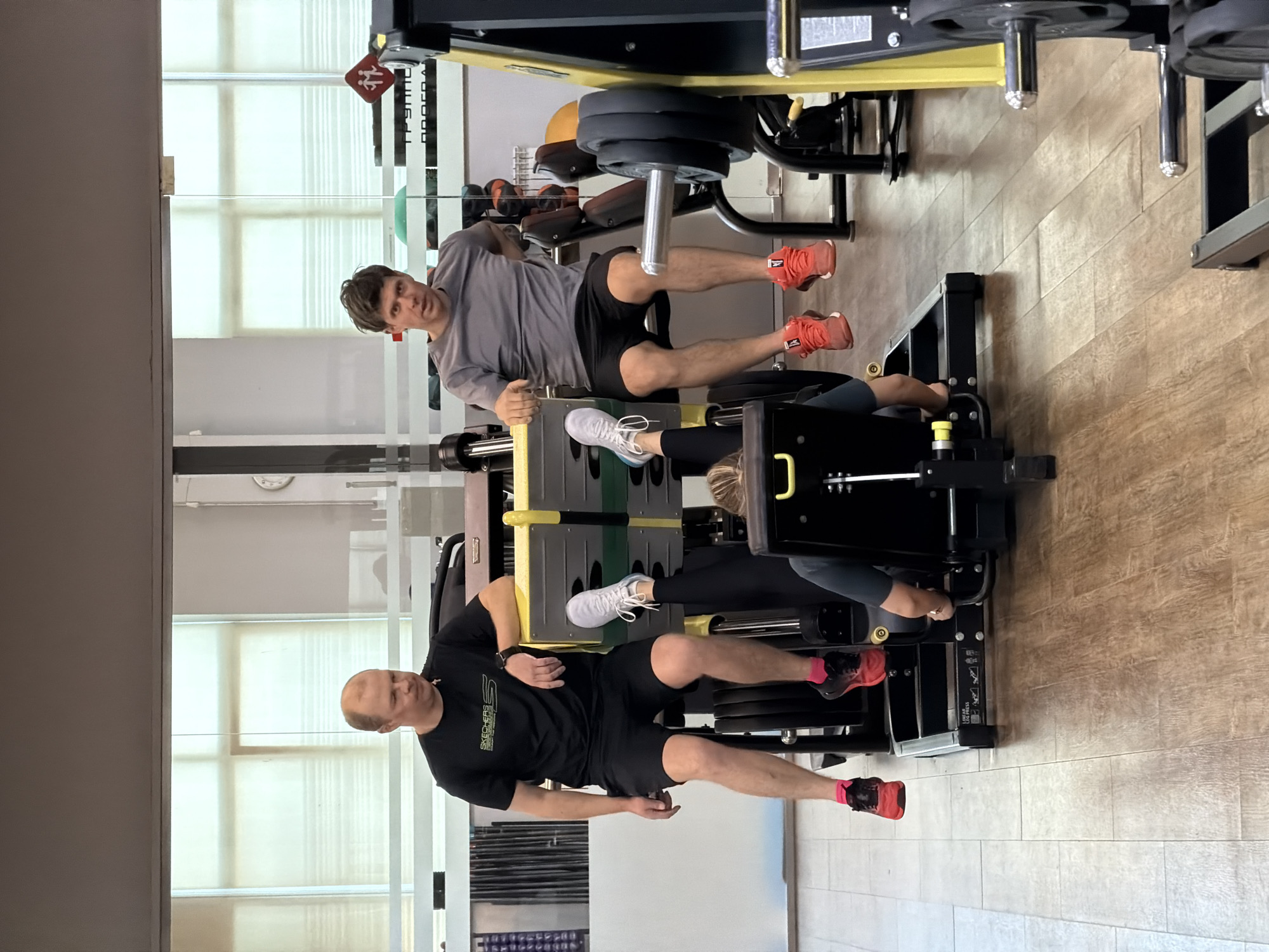Two men are using a leg press machine inside a gym. The gym has a modern interior with wood-look flooring, large windows with blinds, and various weight machines. One man is wearing a grey long-sleeved shirt and black shorts, while the other is wearing a black t-shirt and black shorts with red socks. Both are positioned on the leg press. The city of Chaikovsky, Russia is not discernable.
