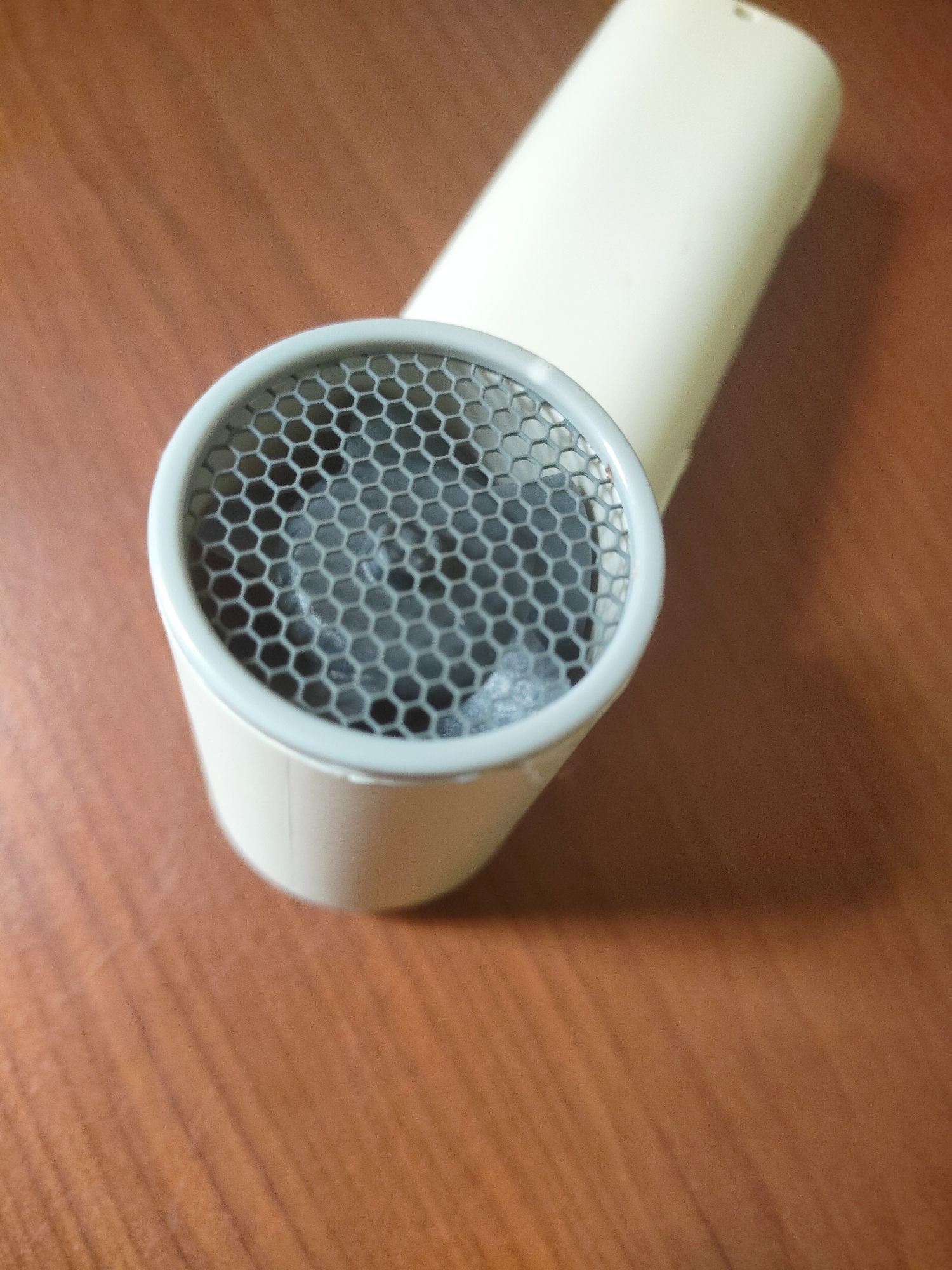 The media file is a close-up, high-angle shot of a beige hairdryer resting on a brown wooden surface. The hairdryer's nozzle, which has a hexagonal grille, is prominently featured in the foreground. The grille is made of a gray material and covers the opening of the nozzle. Inside the nozzle, some blurred dark elements are visible, possibly the fan or heating elements of the hairdryer. The handle of the hairdryer extends upwards and to the right, out of the frame. The wooden surface exhibits a visible grain pattern, suggesting it might be a desk or tabletop. The lighting in the image creates a shadow to the right of the hairdryer, indicating a light source from the left. The overall scene is static, with no activity taking place. No text is visible in the image. The context is unclear, but the composition and focus suggest an emphasis on the object itself.