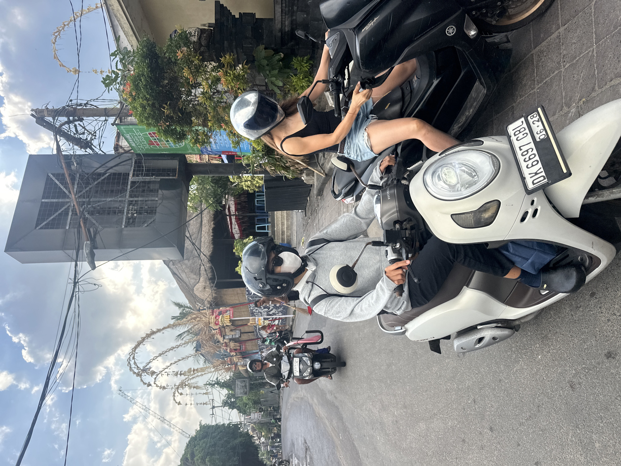 In North Kuta, Indonesia, two people are riding motorcycles down a paved street. In the foreground, a person wearing a gray hoodie, black pants, and black shoes rides a white scooter. They are wearing a white face mask and a black helmet. Beside them, a person with long brown hair wearing a black tank top and denim shorts rides a black motorcycle. They are wearing a silver helmet. The street is lined with buildings with thatched roofs, shops, and trees. Decorative arches made of natural materials are visible along the roadside. Power lines are strung across the sky. A sign for "CANGGU MARKET" is visible on a green banner. The sky is cloudy with patches of blue.