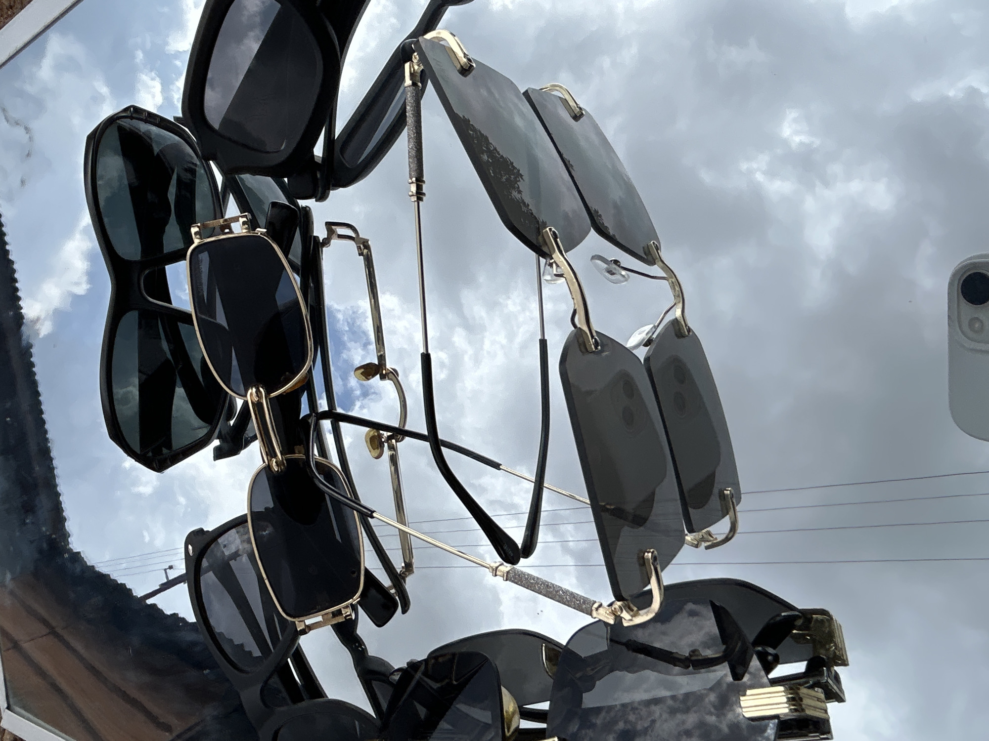 The image shows a collection of sunglasses arranged on a reflective surface, likely a mirror. The sunglasses are of various styles and colors, including black frames with dark lenses and gold accents. The reflective surface creates a distorted image of the sky, which appears overcast with clouds. The scene is set outdoors, possibly in a courtyard or a balcony, in the city of Jos, Nigeria. The overall impression is one of casual luxury and outdoor living.
