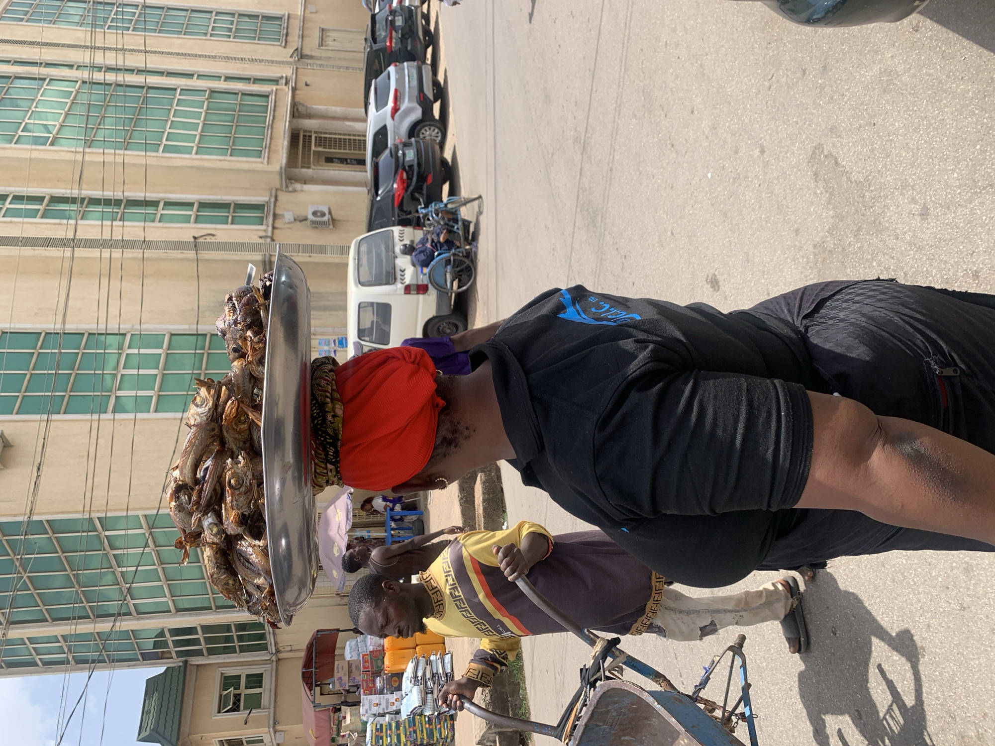 The image depicts a street scene in Jos, Nigeria, showing a woman with a large metal tray of what appears to be dried fish balanced on her head. She is wearing a red headscarf and a black t-shirt. A man is next to her, pushing a wheelbarrow. Behind them, there is a building with multiple windows, several parked cars, and various shops and street vendors.