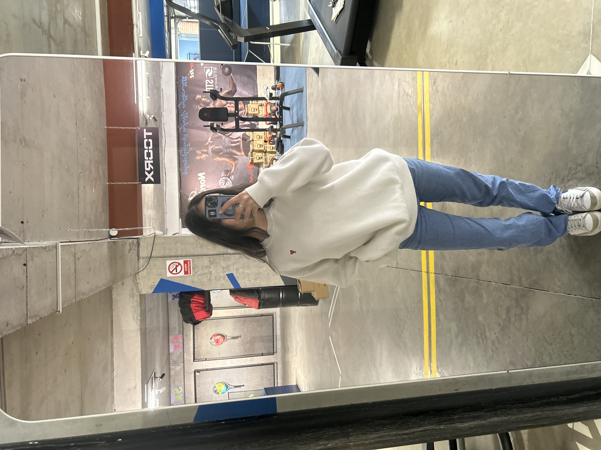 A person is taking a mirror selfie inside a gym. The gym features exercise equipment and advertisements in the background, and tennis rackets in metal cages. A sign that says "XCOOT" can be seen, as well as advertisements written in Georgian. The city is Tbilisi, Georgia.