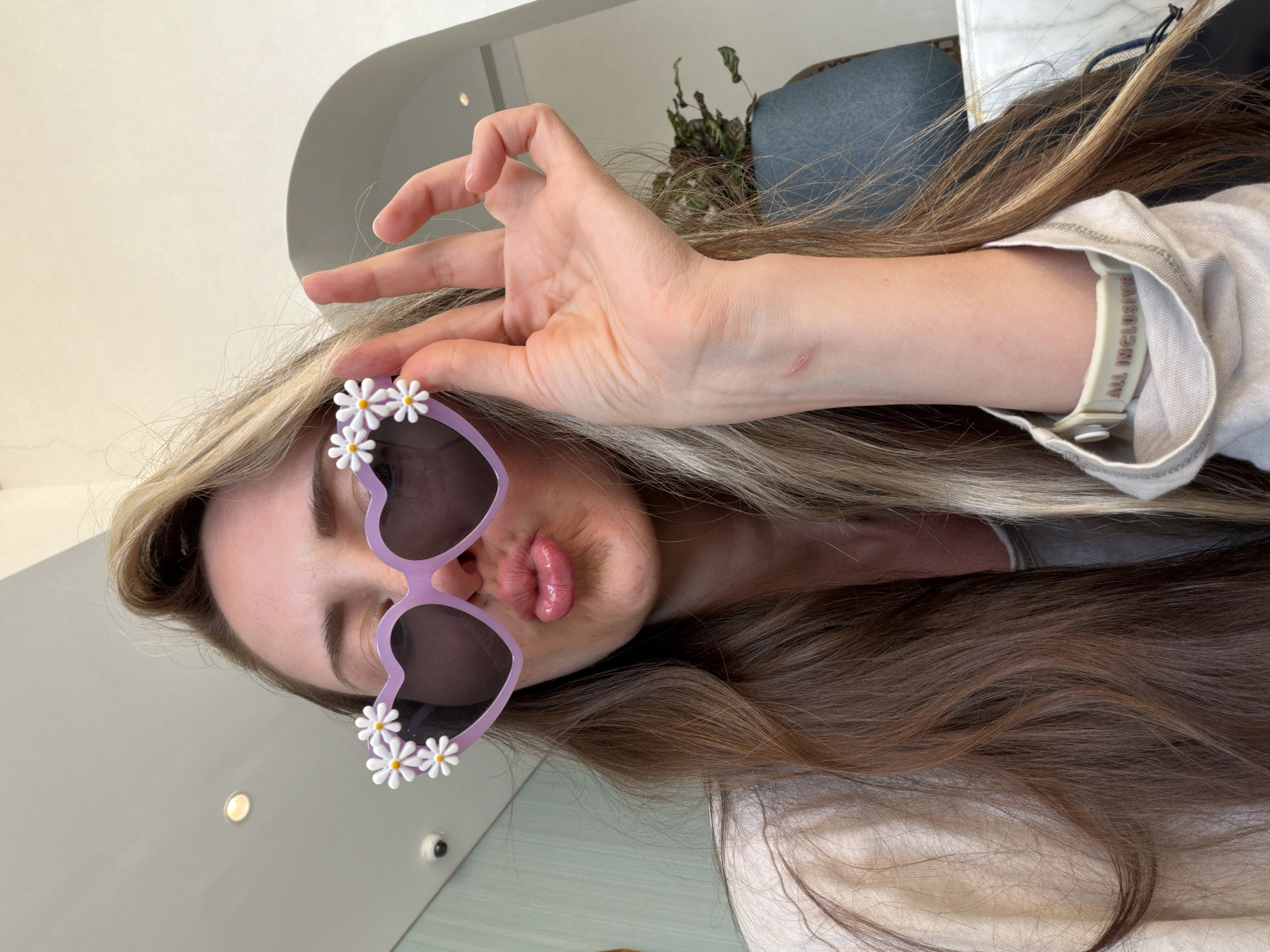 The image captures a person, likely female, posing playfully with her head tilted to the right. She is wearing distinctive lavender, heart-shaped sunglasses adorned with white daisy flowers on the lower frames, covering her eyes. Her lips are visible below the sunglasses, forming a pouty or kissing expression.
Her right hand is raised to her head, making an "OK" gesture with the thumb and index finger forming a circle, and the other three fingers extended upwards. Long, wavy hair with blonde highlights flows around her and across the frame. She is wearing a light-colored, possibly white or off-white, short-sleeved shirt. A white wristband is visible on her right wrist, partially covered by the shirt sleeve, with the words "ALL INCLUSIVE" clearly printed in a reddish-brown color, suggesting a resort stay. A small, faded scar is visible on her forearm, above the wristband.
The background is a bright indoor setting, featuring a light-colored wall or ceiling in the upper left, and hints of a dark blue upholstered item (possibly a chair or couch) and a green potted plant in the upper right. The bright lighting indicates it is daytime. The overall impression is one of a carefree, playful moment, consistent with a vacation in Sharm El Sheikh, Egypt.